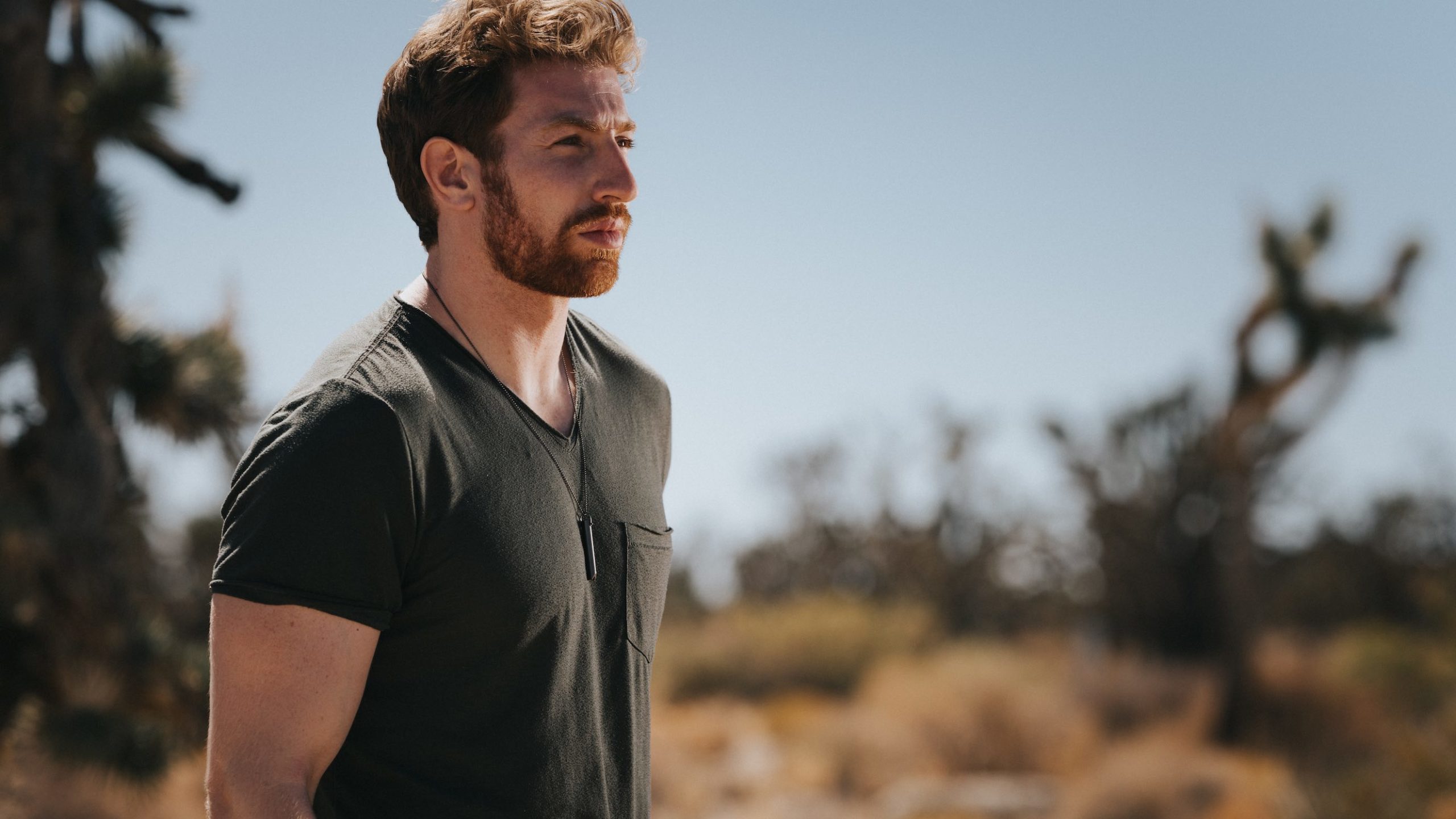 Muscular male with short, red hair and beard wearing a black tshirt looking off into the distance wearing a Komuso whistle around his neck
