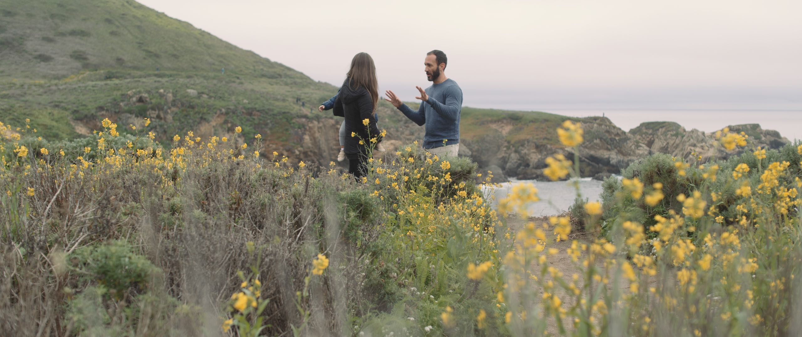 Dailies 203 Big Sur Video Production Company Los Angeles Still showing man and woman who is holding a child talking to each other near the coastline with wildflowers in the foreground