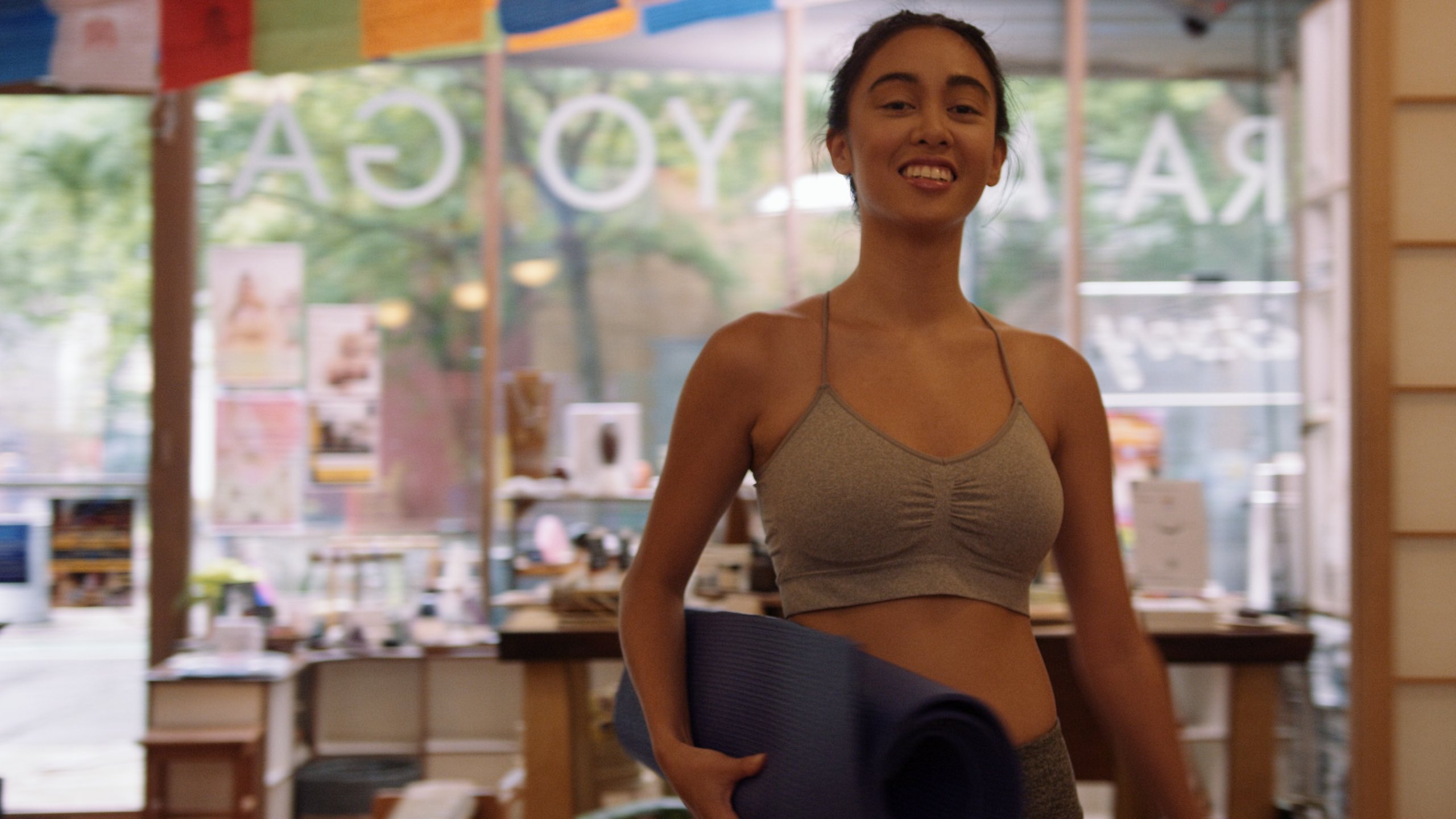 Apples & Oranges Woman wearing a gray sports bra top holding a yoga mat smiling for the camera