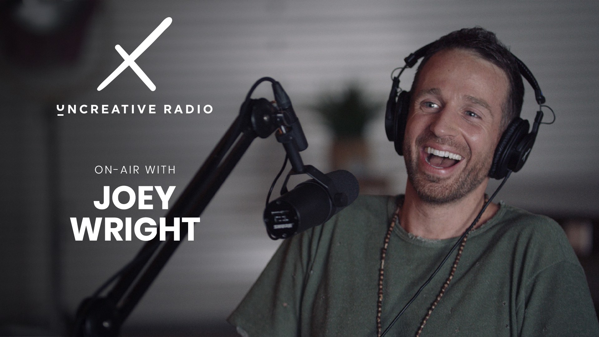 White X Uncreative Radio logo on air with Joey Wright title with him with short beard and hair wearing black headphones and olive green shirt smiling for the camera by a microphone