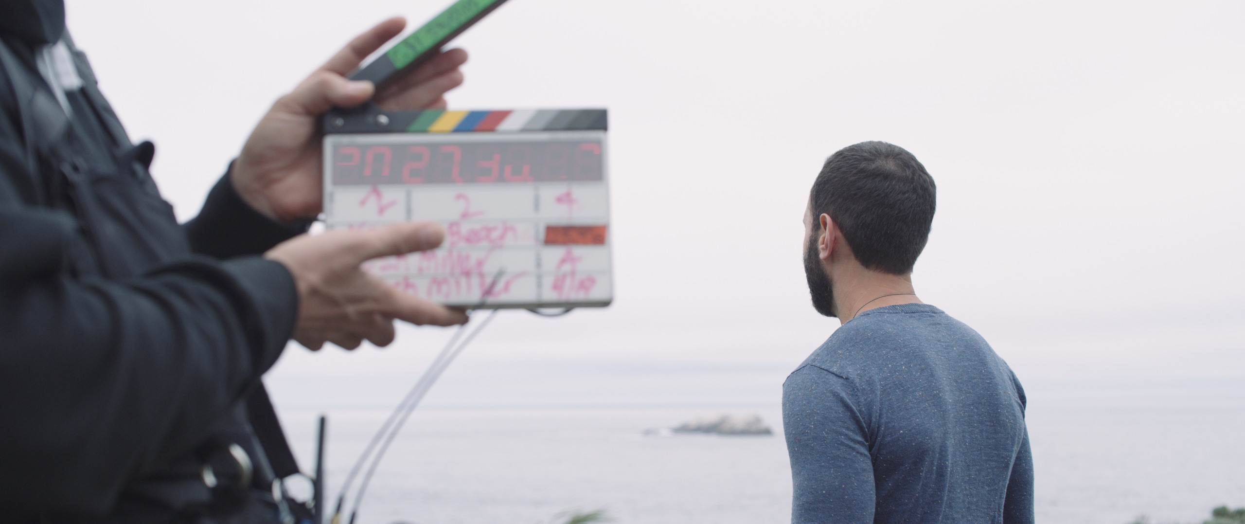 The Dailies Episode 3 BTS Footage for Komuso with view from behind of man wearing a blue shirt looking out over the water with a crew member holding a clapboard behind him