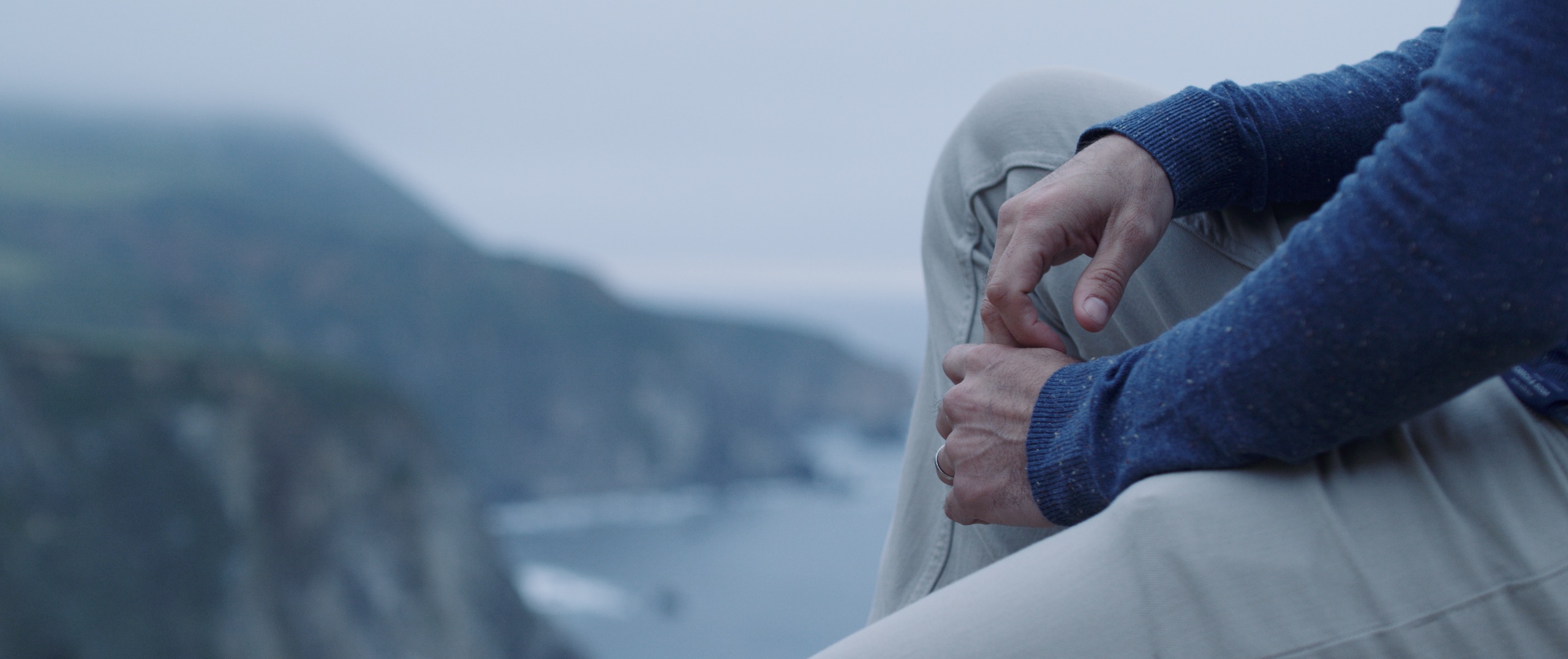 The Dailies Season 2 Episode 3 Komuso Big Sur with closeup of man wearing a blue sweater and gray jeans sitting by cliffs with water down below
