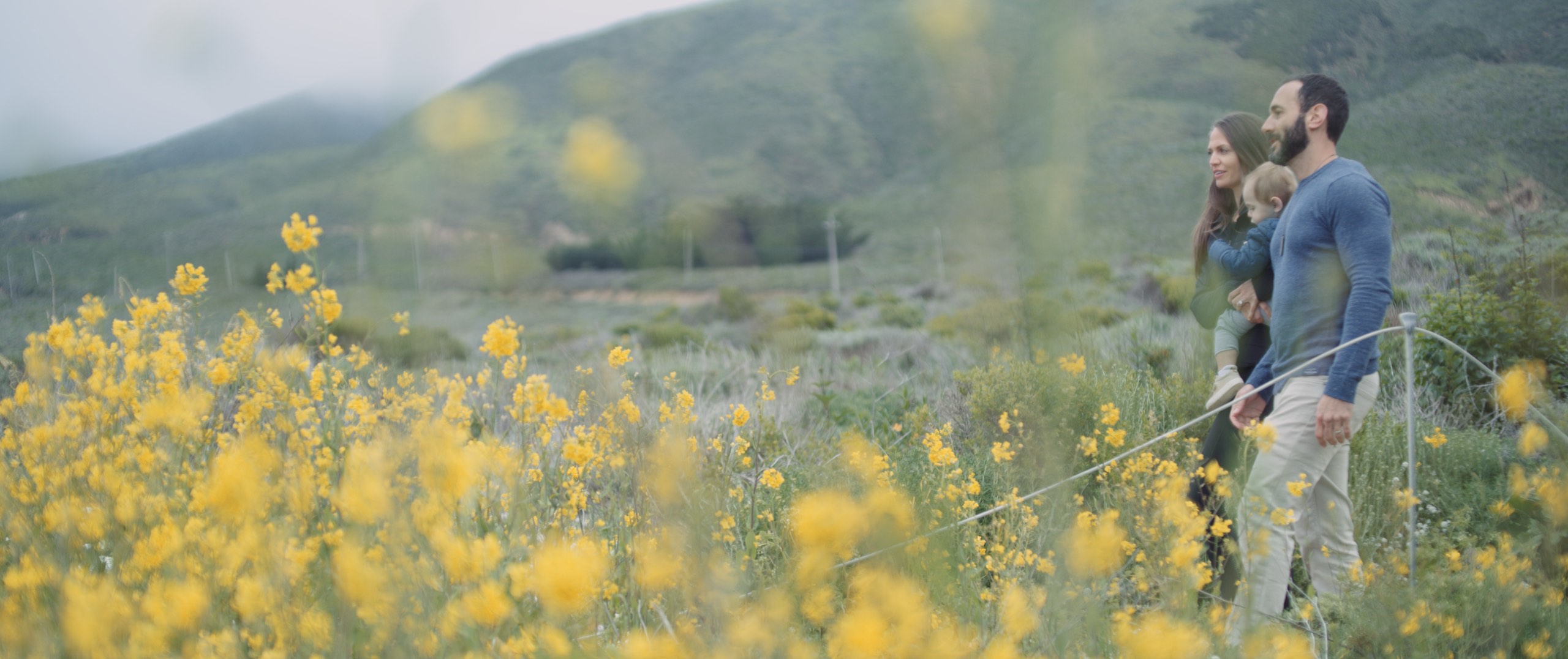 The Dailies Season 2 Episode 3 Komuso Big Sur with side profile of man and woman carrying child walking in the fields with yellow wildflowers