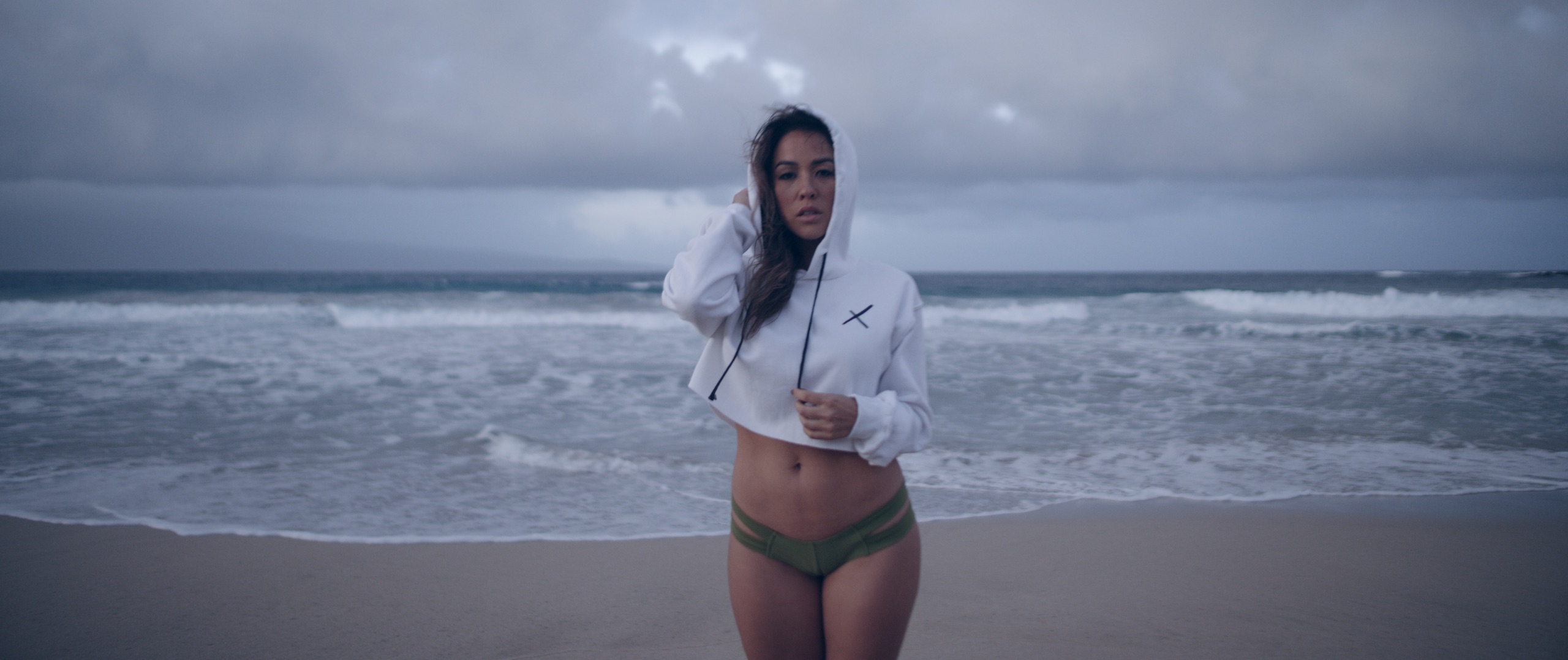 The Dailies Episode 4 The Making of Uncreative Merchandise Shop Female model posing wearing white cropped Uncreative sweatshirt and olive green thong standing on a beach by water
