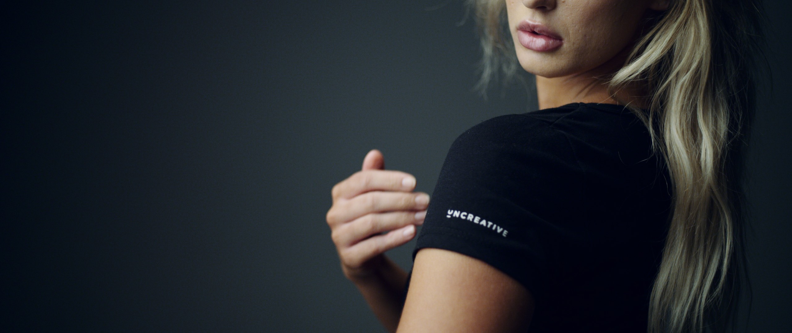 The Dailies Episode 4 The Making of Uncreative Merchandise Shop Side profile of woman with long blond hair looking over her shoulder wearing a black Uncreative t shirt showing the Uncreative logo posing for the camera