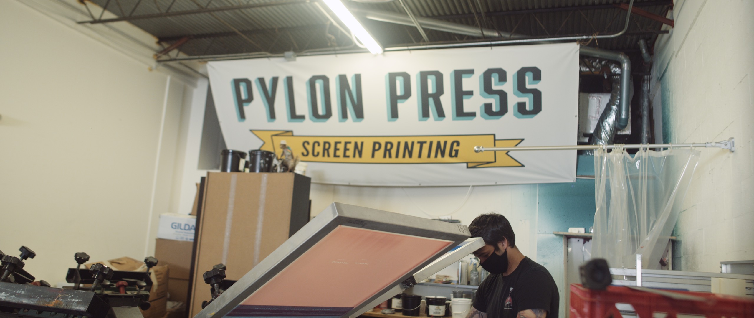 The Dailies Episode 4 the Making of Uncreative Shop and Merchandise with man wearing a black mask working with equipment in Pylon Press Screen Printing Shop