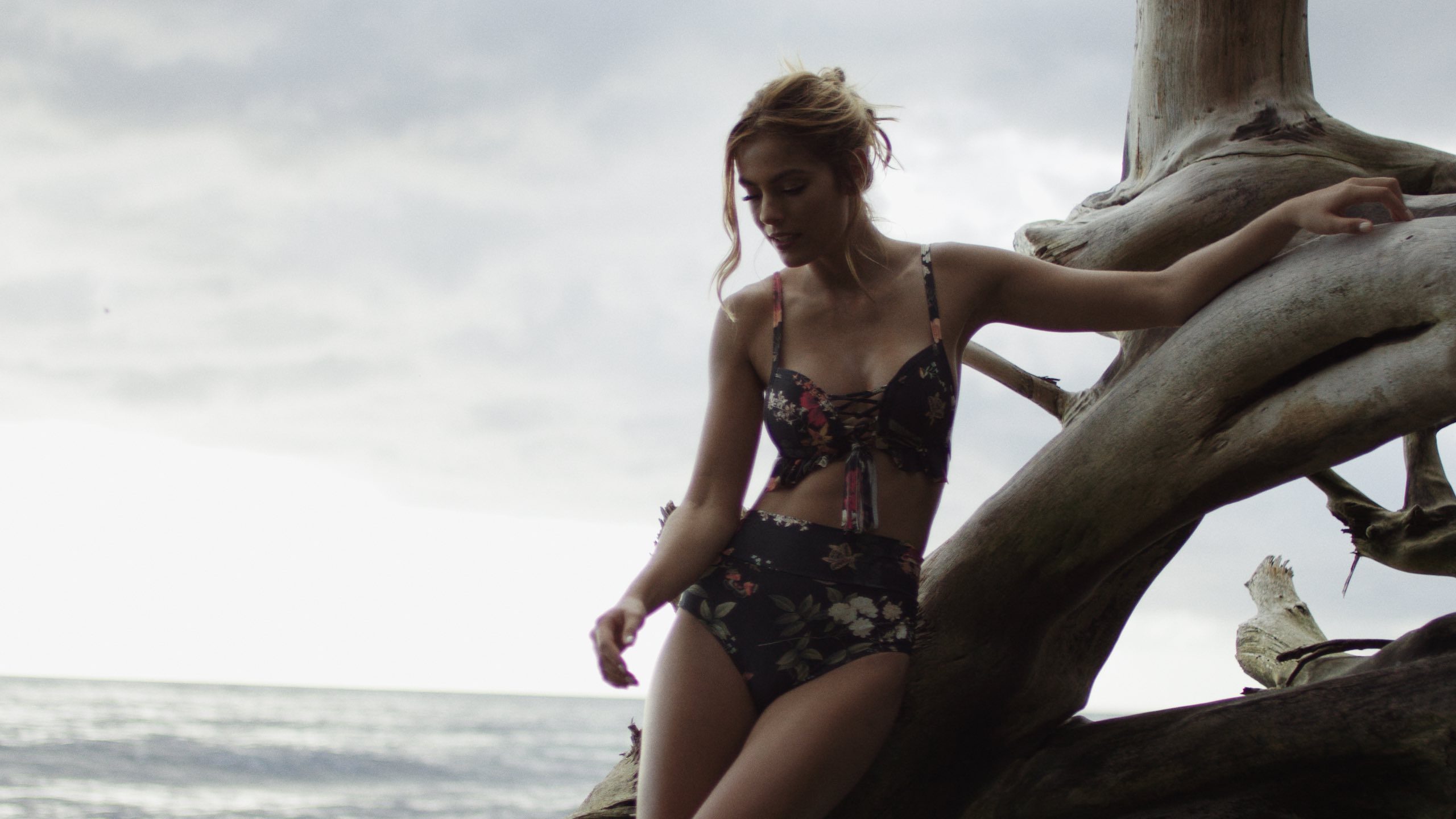 Puerto Rico with Montce Swim, The Dailies Season 2 Episode 1 with closeup of model with long blond hair posing wearing dark flowery bathing suit leaning on a dead tree on a beach