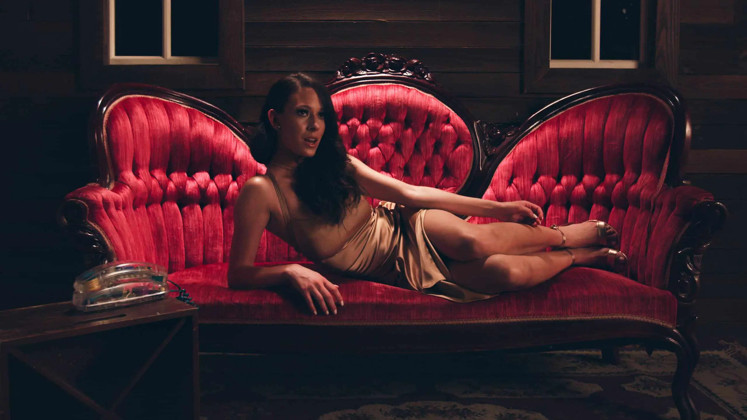 Sabela Love Jones posing on a vintage red velvet lounge chair wearing gold colored dress and high heels with a see through landline phone on a table nearby