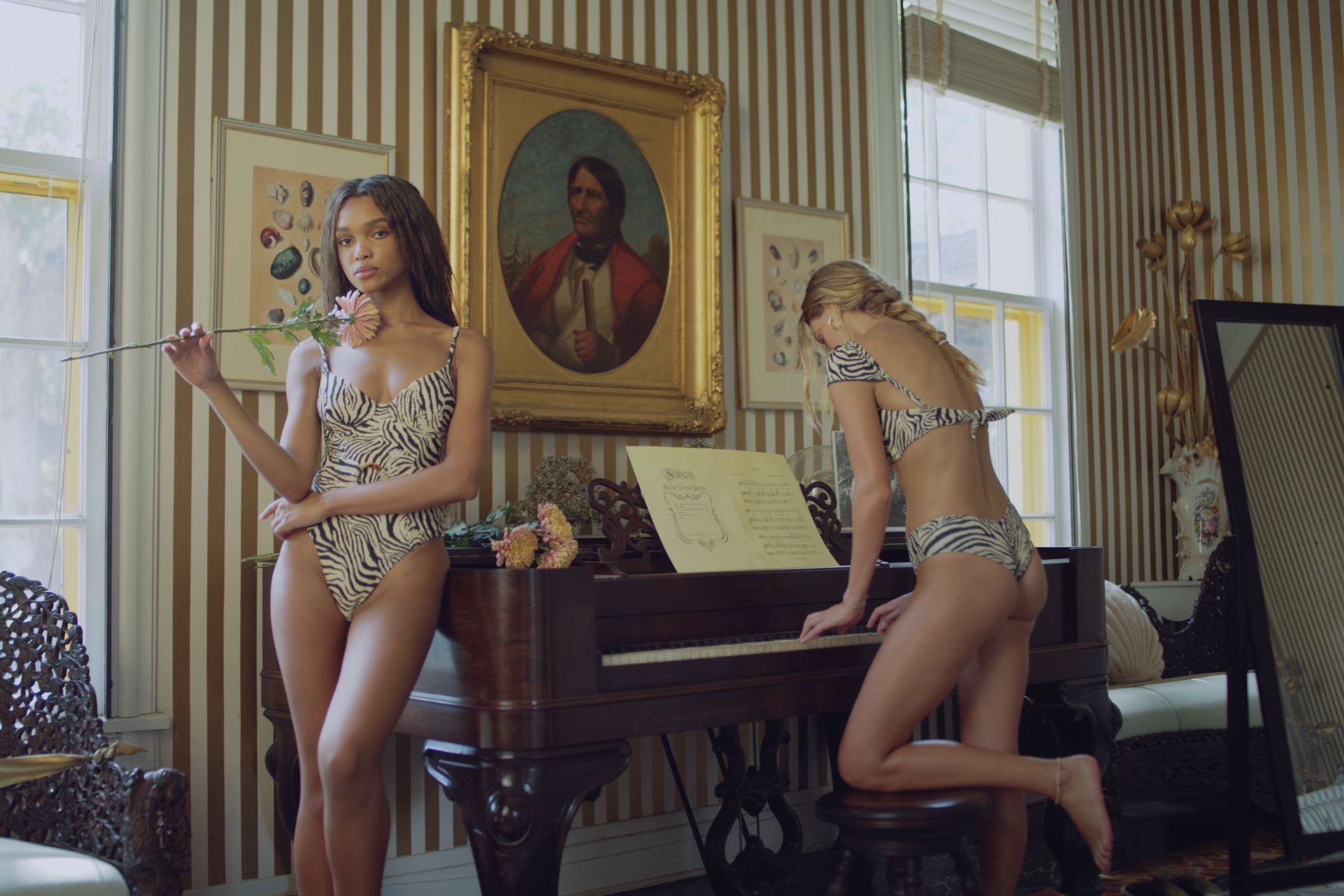 Production Resources Location with two women wearing black and white patterned swimsuits with one leaning against the piano holsing a flower and the other playing the piano