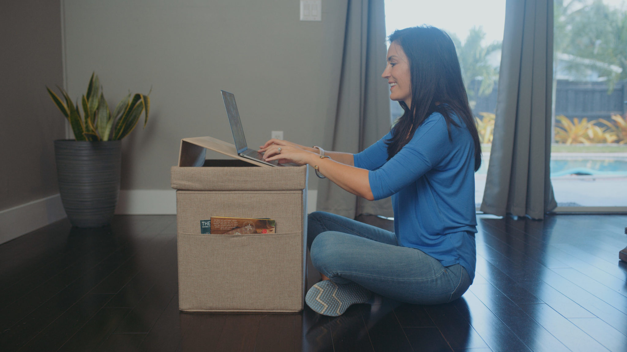 Audy Global Enterprise Ottoman Laptop Feature Fort Lauderdale Florida C&I Studios Side Profile Of Woman Wearing Blue Top And Jeans Sitting On The Floor Going Through A Box