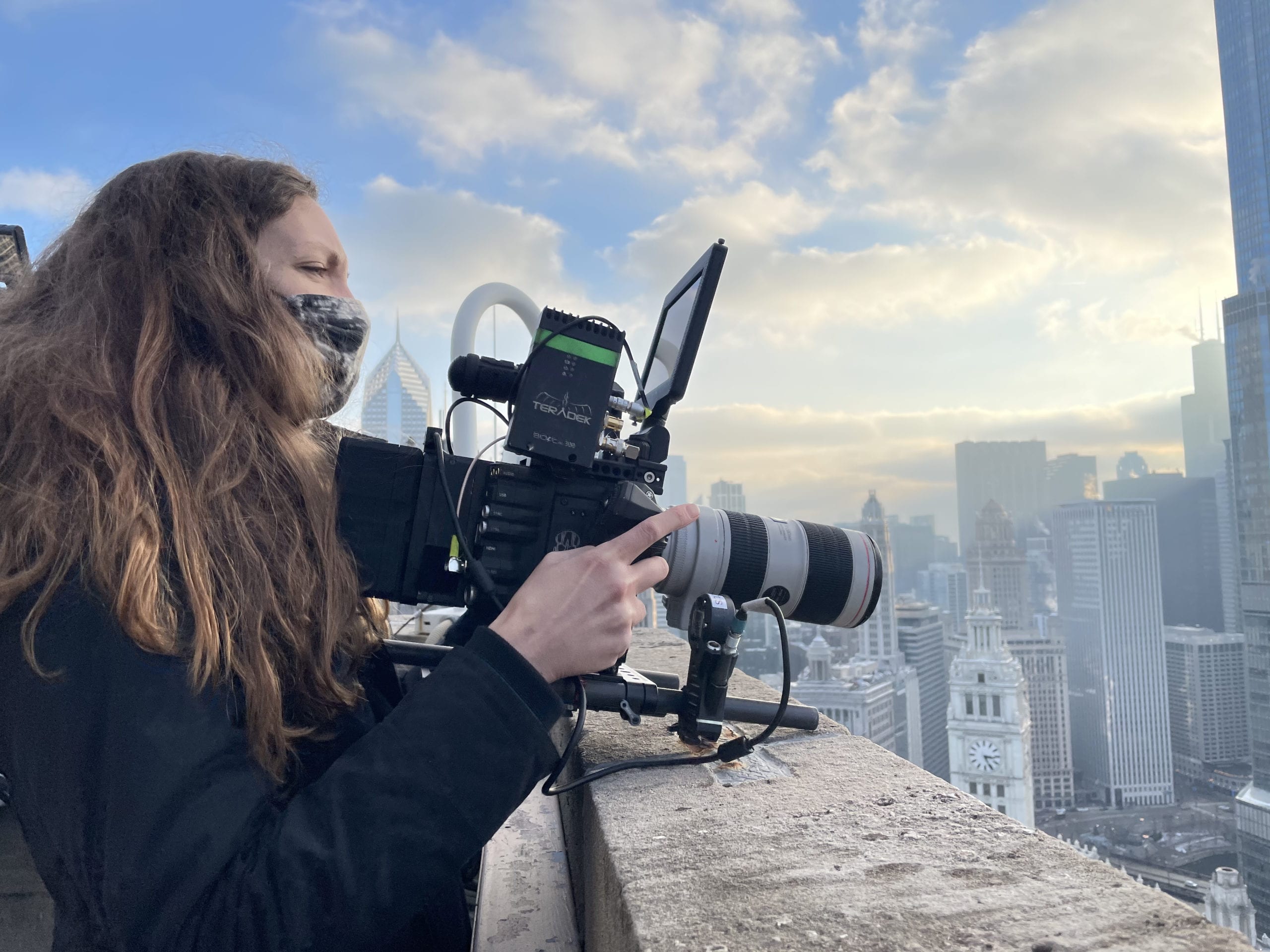 Side profile of female crew member with long blond hair wearing a black mask using a camera on a rooftop in a city