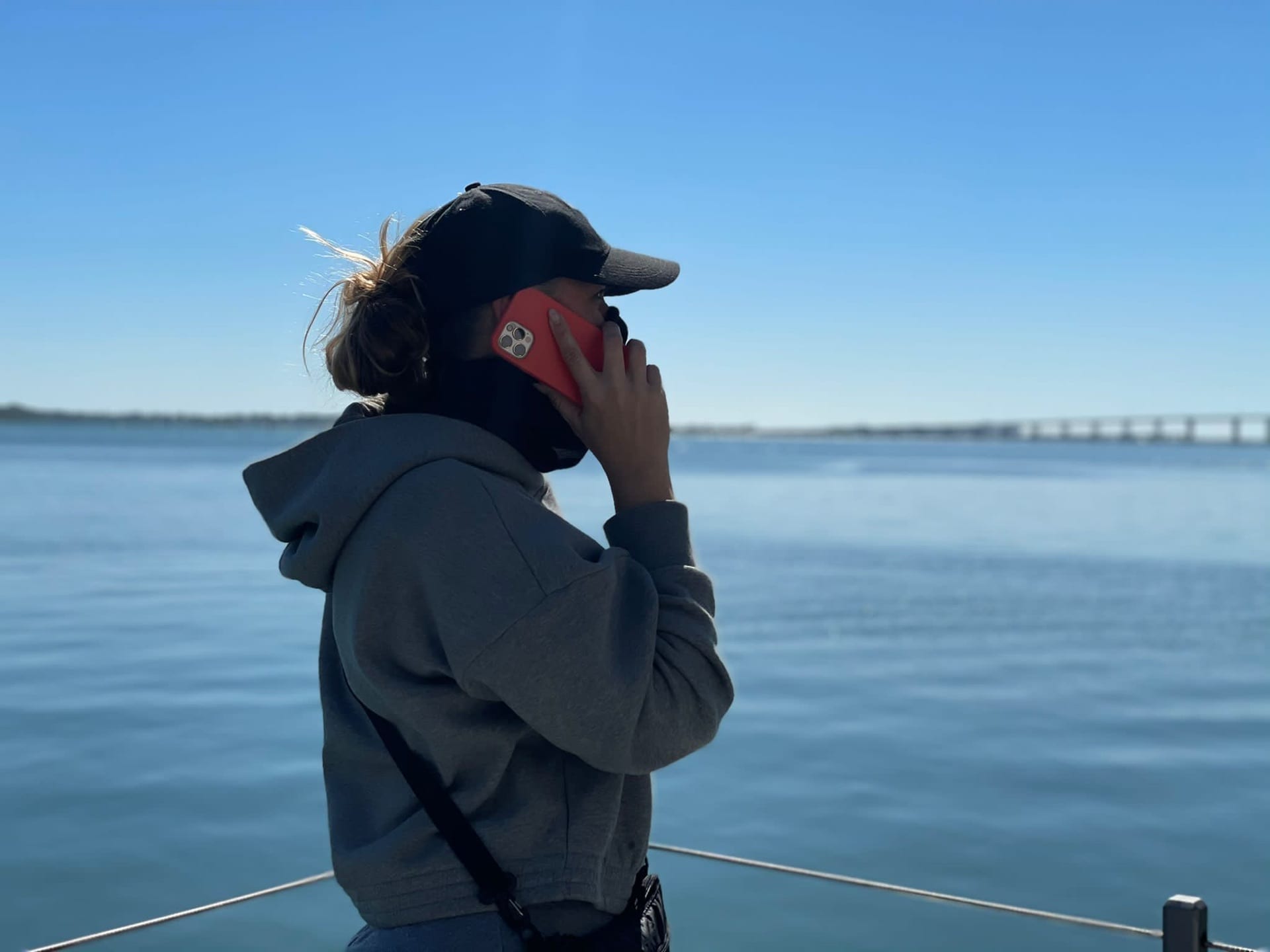 Side profile of woman wearing a black cap and gray sweatshirt talking on a dark orange cell phone