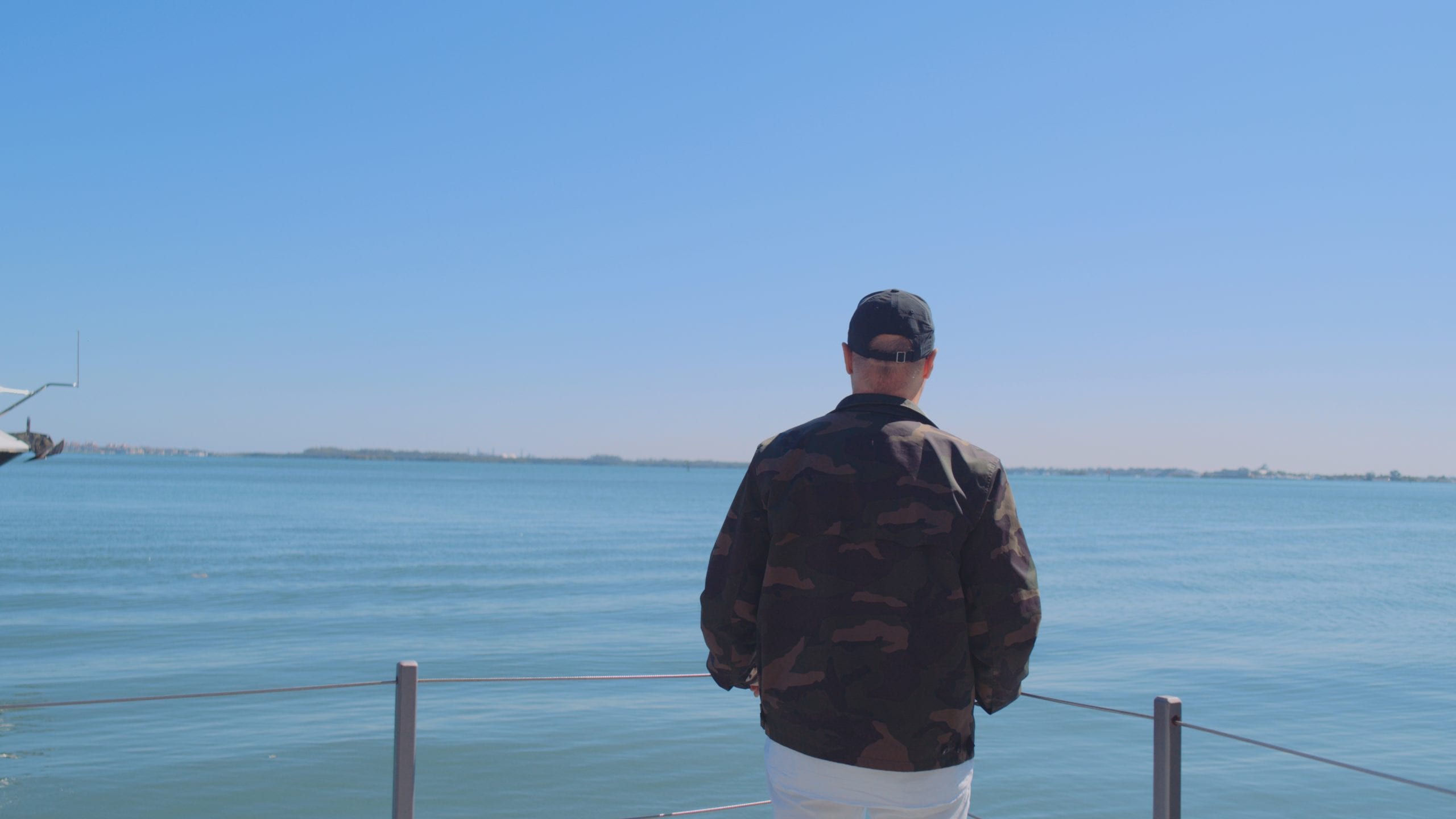 View from behind of man wearing a cap and camouflage jacket standing on a balcony looking out over the water