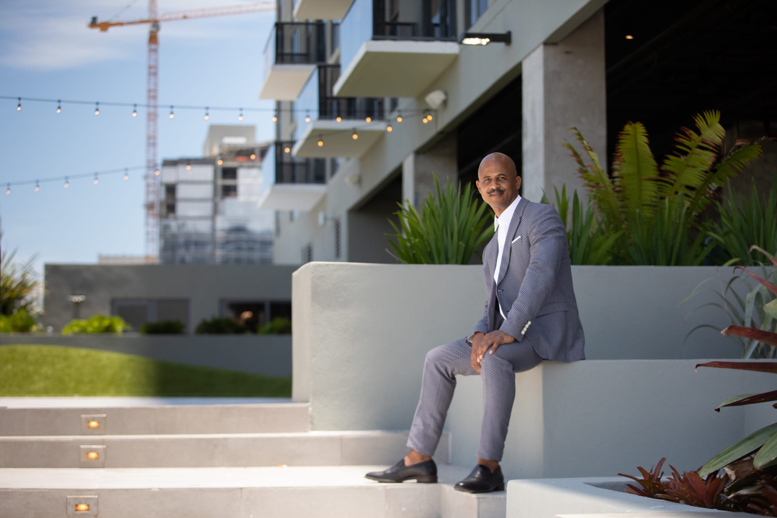 Bald African American male model with moustache wearing gray suit and white shirt sitting near stairs