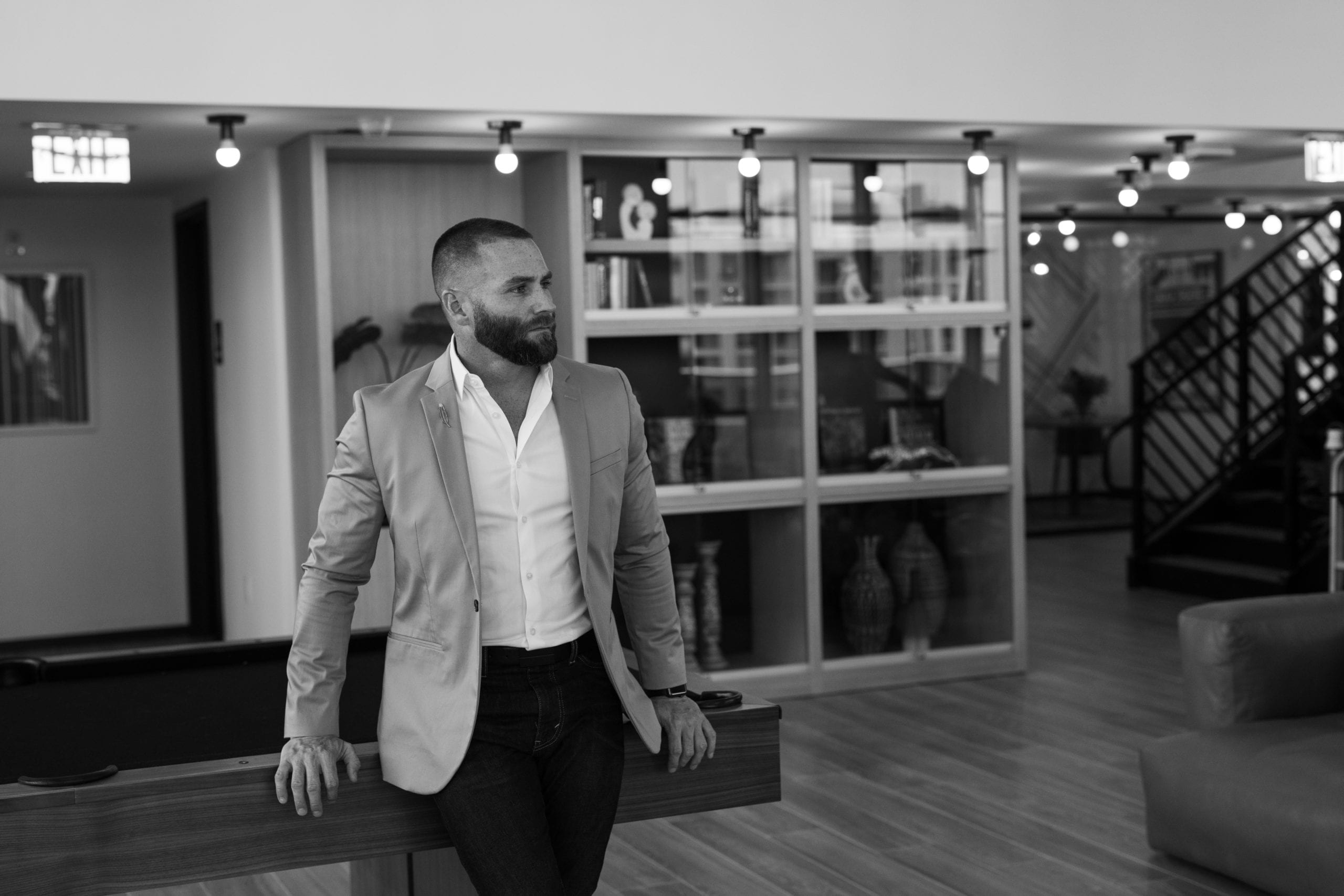 Black and white of male model with beard in a suit leaning back on a pool table looking off to the side