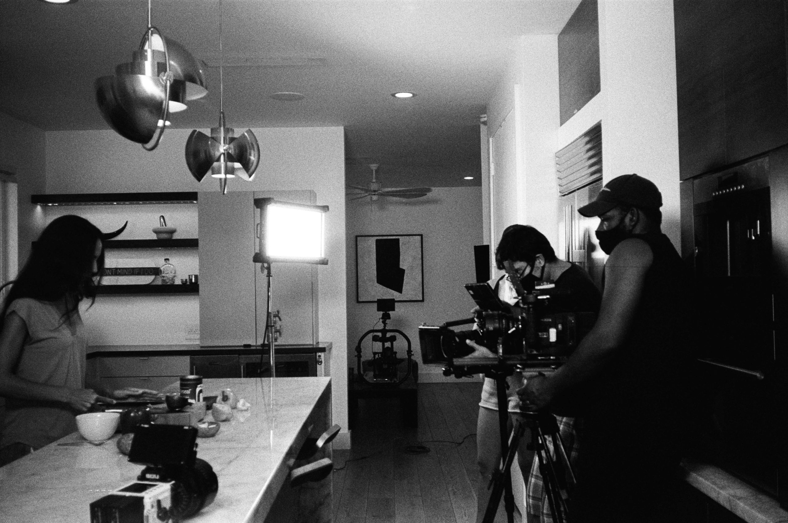 Promotional Video for My Business. Black and white photo of a cooking show in progress with a woman cooking at the counter and two males wearing masks filming the show. There is another video camera on the counter as well.