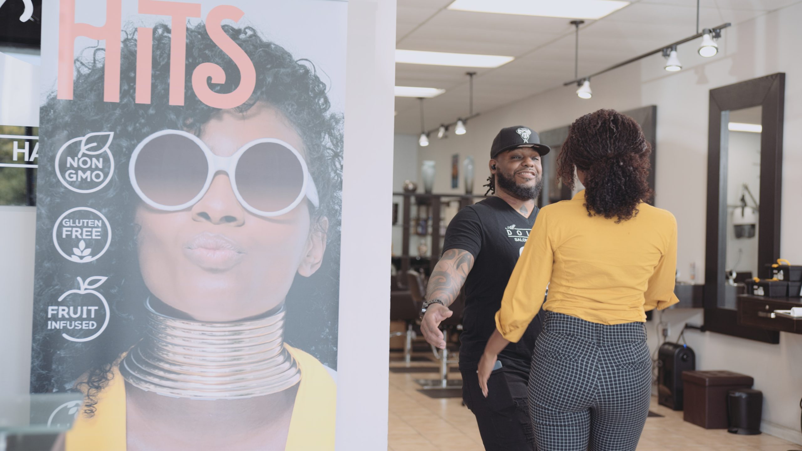 All Dolled Up Month 1 Magazine color and view from behind of woman wearing a yellow top and black and white pants with fizzy hair greeting a male hairdresser at a salon