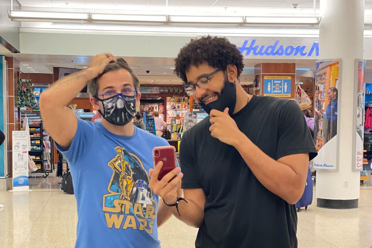 Crew member with mask down showing cell phone to another man wearing a light blue Star Wars t shirt and mask in front of a Hudson News store in a mall