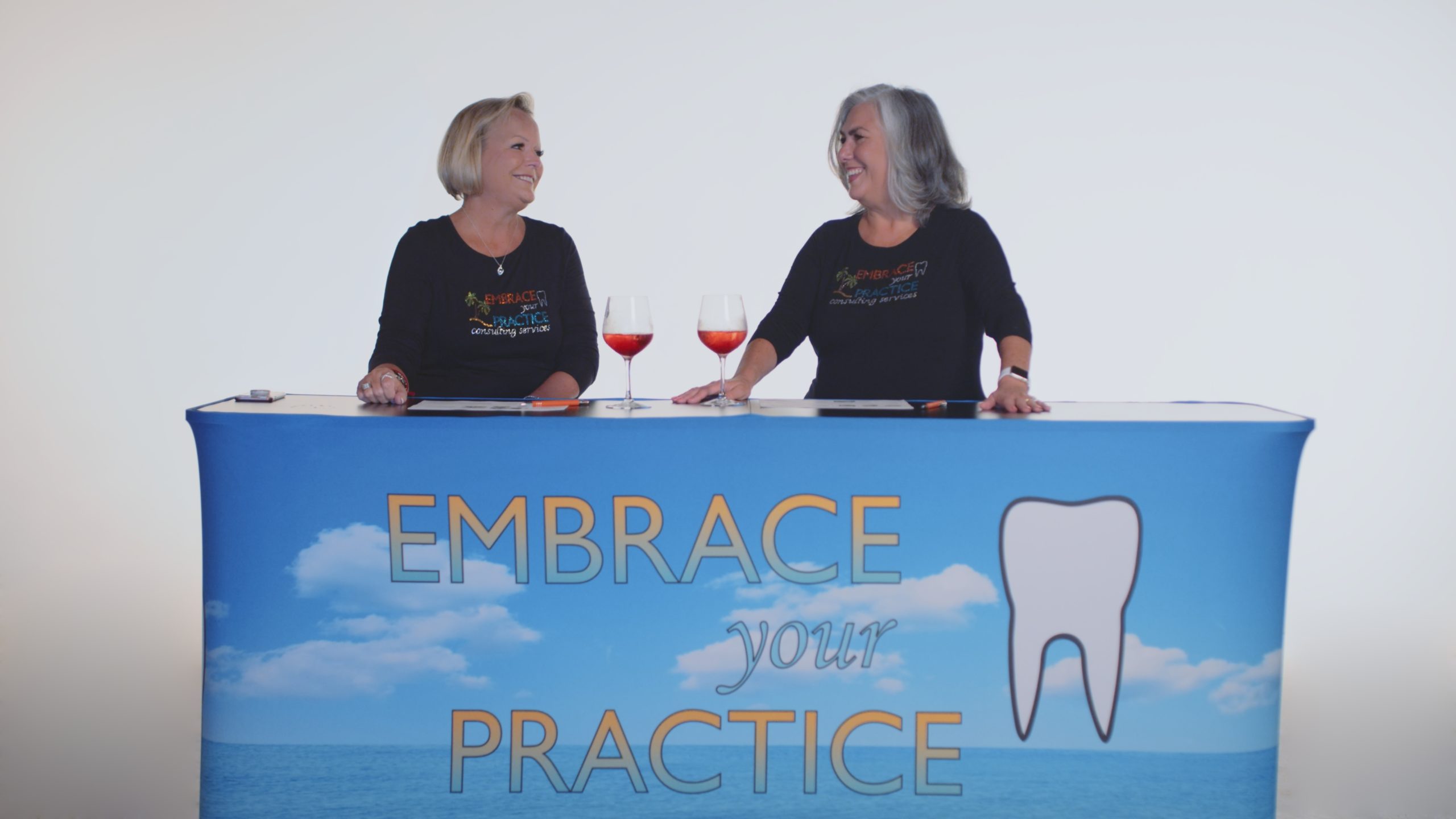 Embrace Your Practice Two women sitting at a table with banner and glasses of wine smiling at each other