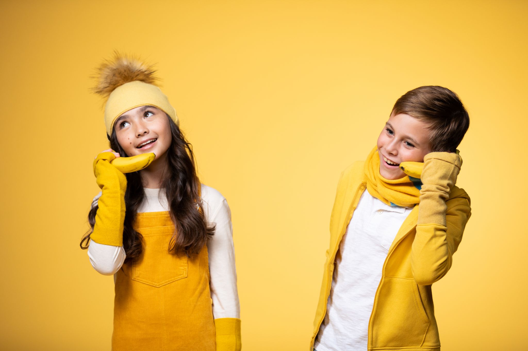 IU C&I Studios Portfolio BB Sheep Lifestyle Colors PROOF Boy model wearing a yellow scarf. jacket and white top and girl model wearing a yellow headband, hand sleeves and overalls as well as white top posing for the camera both using bananas as phones against a yellow backdrop