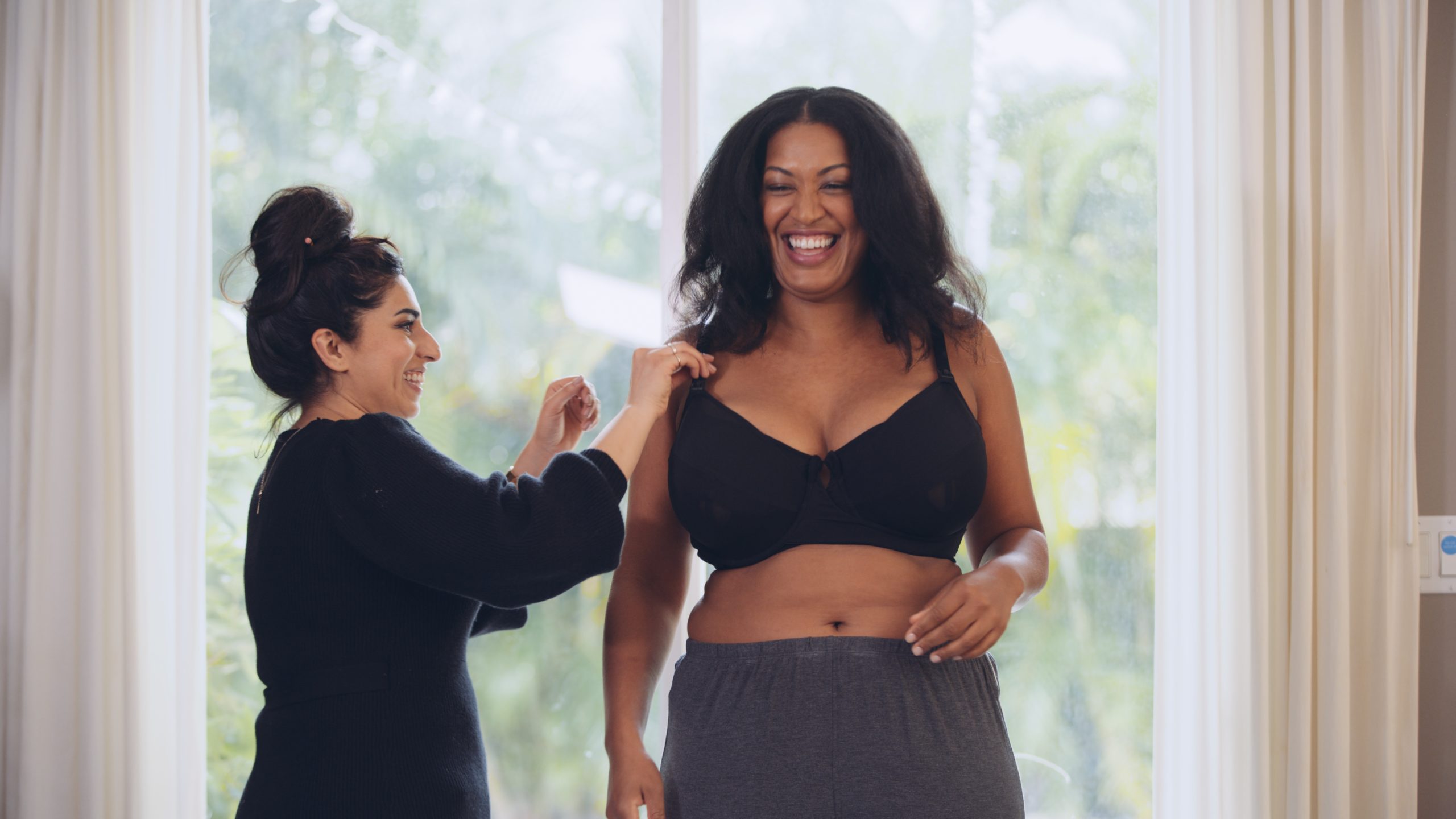 Woman with long hair in a bun adjusting bra strap on model with long brown hair and wearing black bra and grey sweatpants with both of them smiling