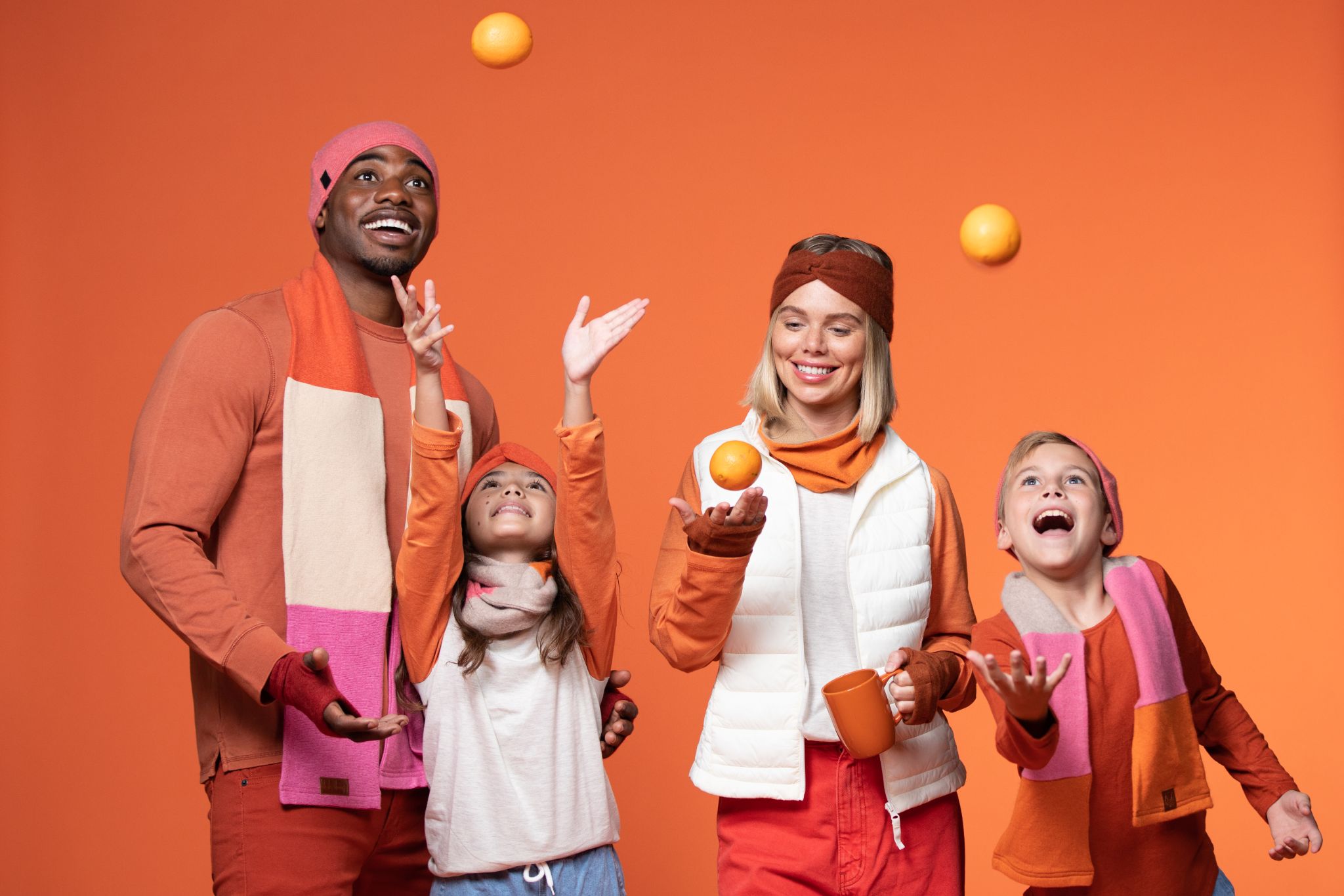 An African American male and a Caucasian woman are juggling oranges with two children against an orange backdrop. They are primarily wearing orange clothing as well.