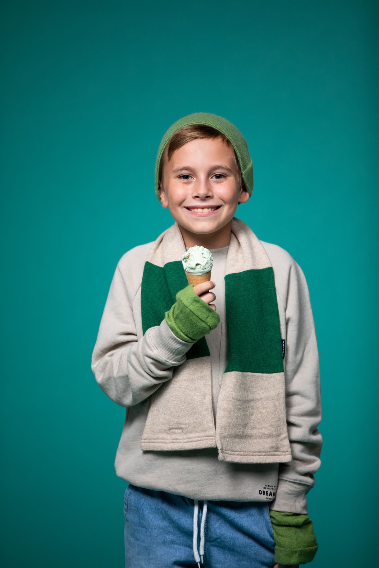Boy with short blond hair against a teal background wearing a gray sweatshirt with blue jeans holding an ice cream cone smiling and posing for the camera.