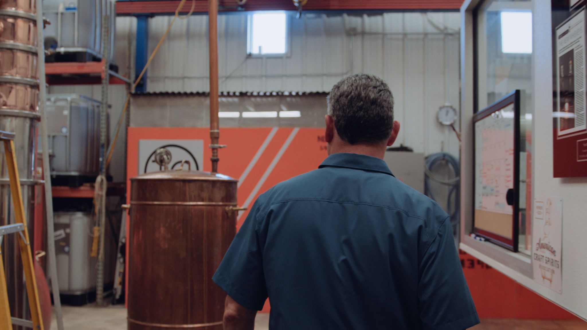 IU C&I Studios Portfolio Taylor Garrett Spirits View from behind of man wearing a blue shirt looking at distillation equipment in a warehouse