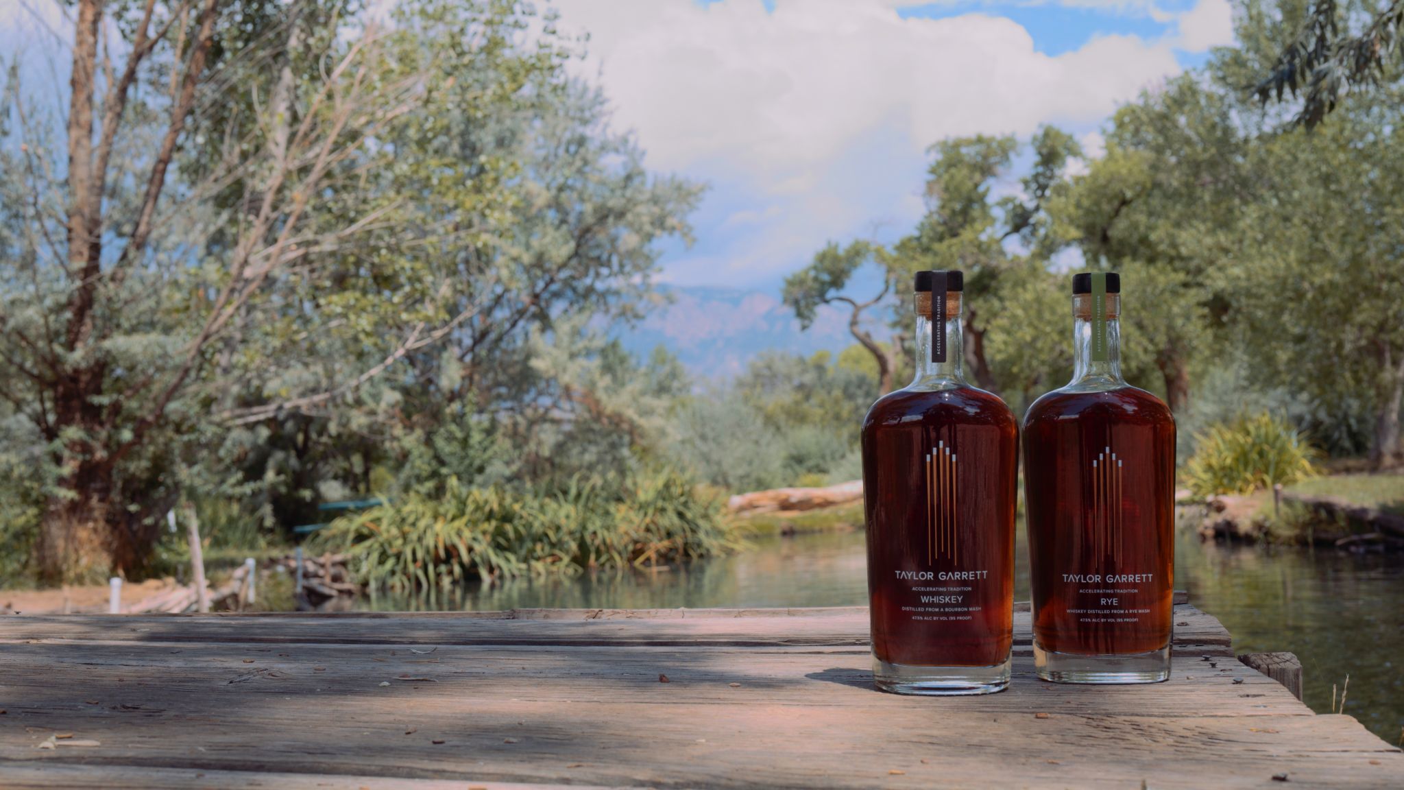 IU C&I Studios Portfolio Taylor Garrett Spirits Two bottles of whiskey on display on a picnic table by a river in the forest