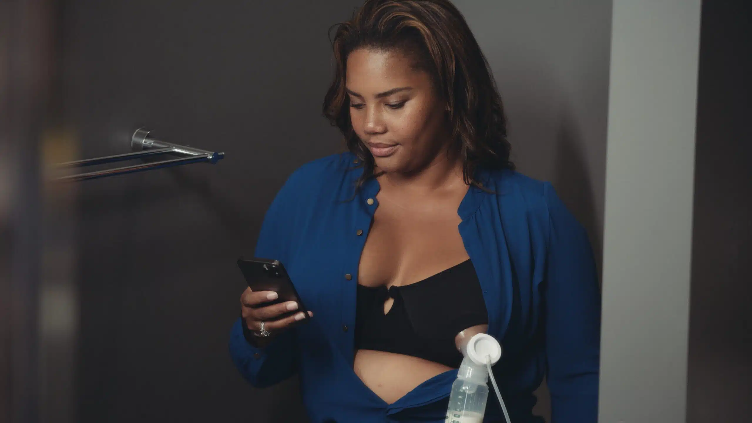 Woman with long brown hair wearing a black bra with breast pump attached and blue shirt looking down at cell phone she is holding