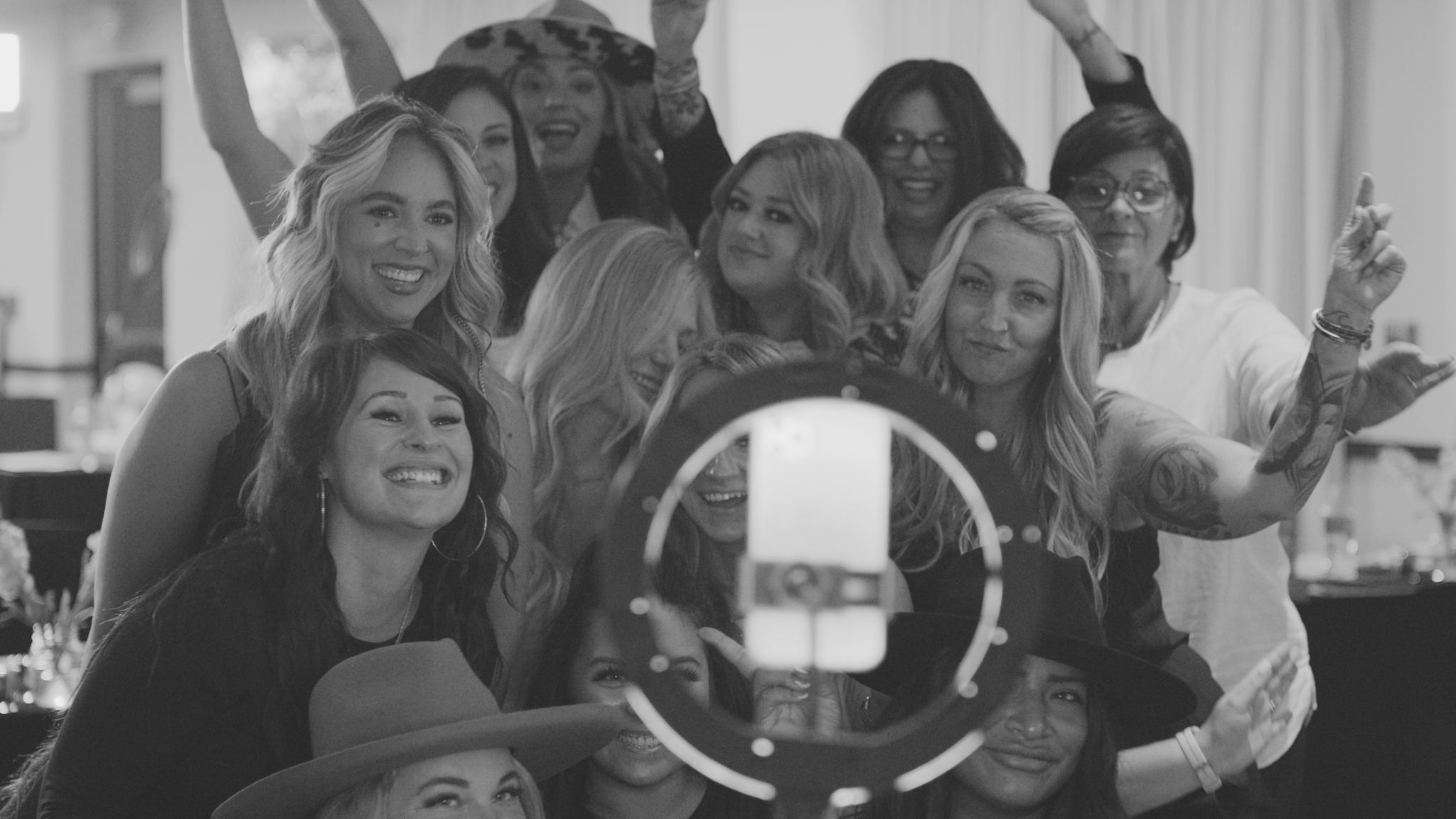 Black and white still of a group of thirteen women posing for the camera with lighting equipment shining on them