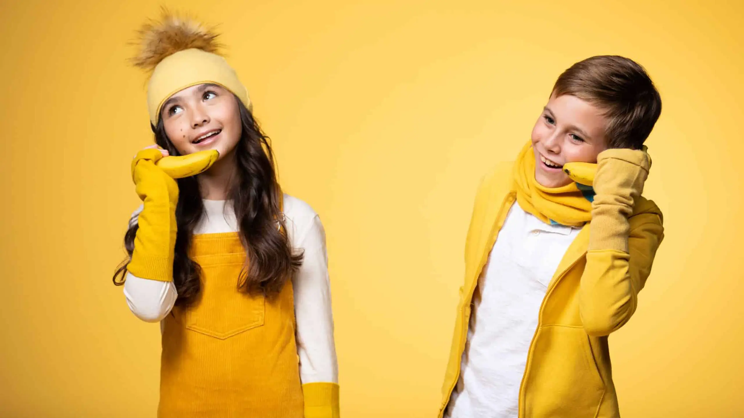 Boy model wearing a yellow scarf. jacket and white top and girl model wearing a yellow headband, hand sleeves and overalls as well as white top posing for the camera both using bananas as phones against a yellow backdrop