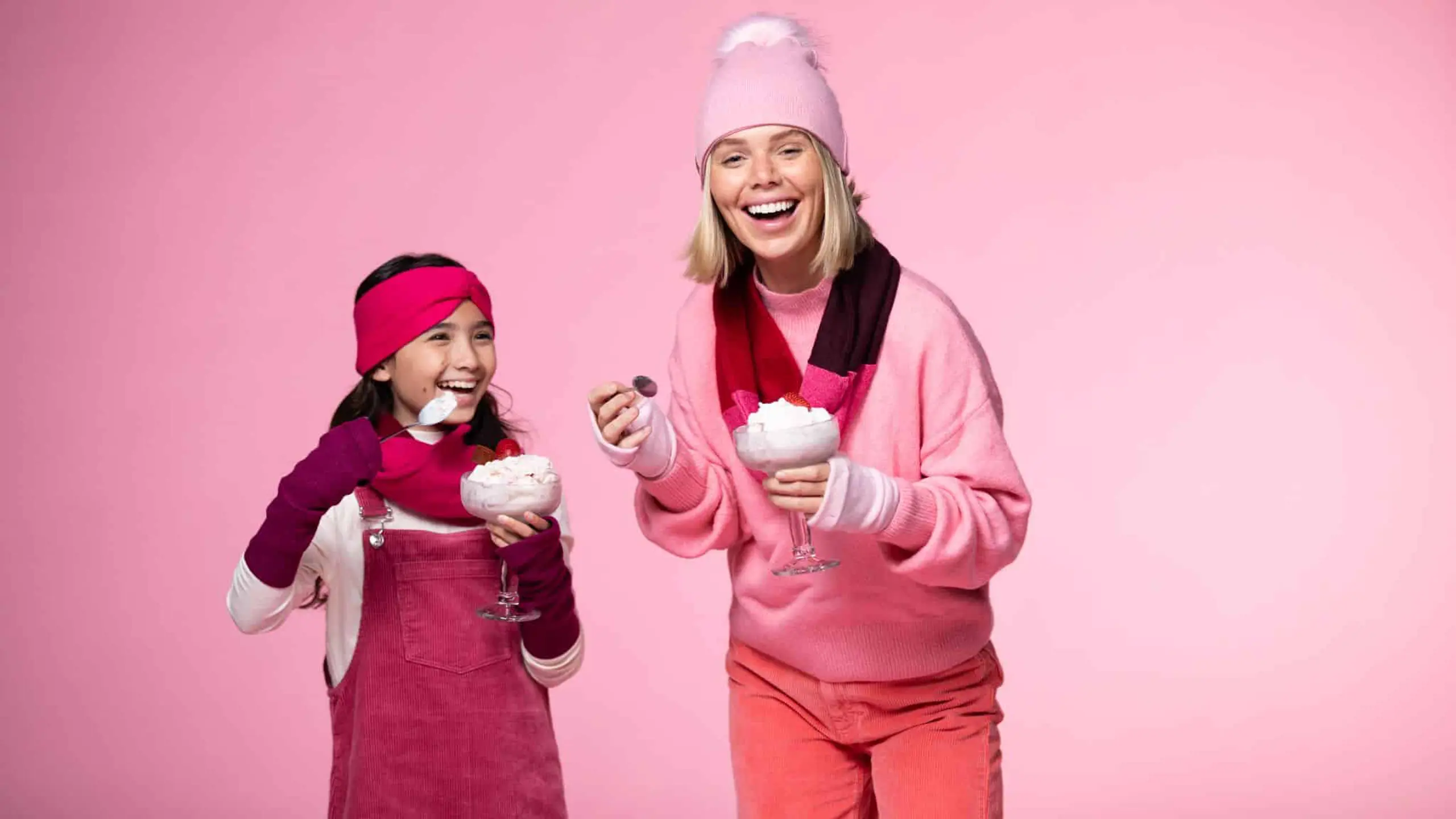 Girl and female models posing for the camera smiling holding ice cream sundaes wearing red and pink outfits respectively against a pink backdrop