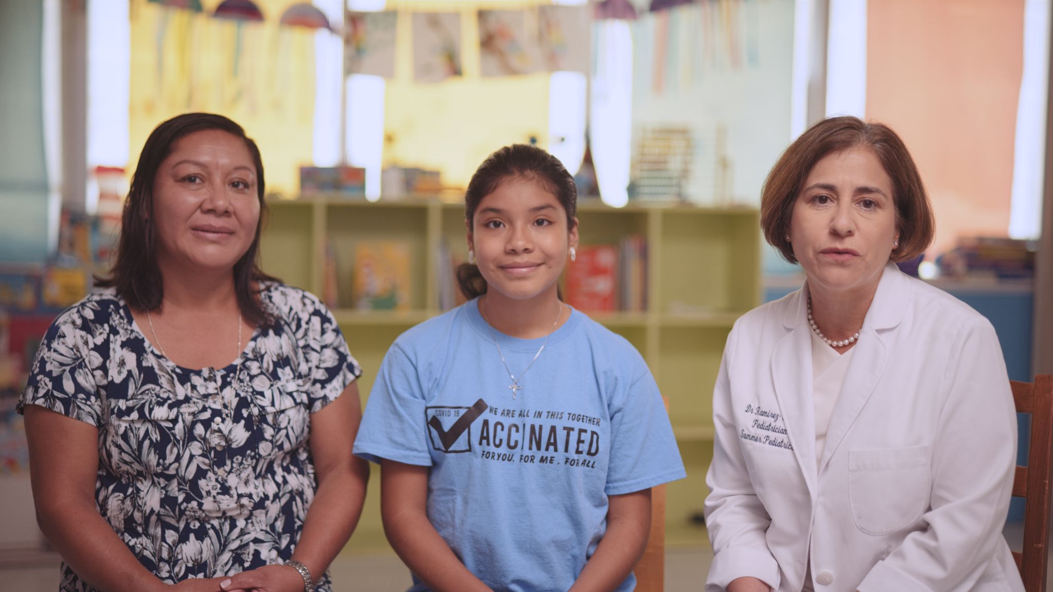 Two female adults and a female youth pose for a picture. Woman on left is wearing a flowery dress, youth in the middle wearing a light blue vaccination t-shirt and female on right is a pediatrician in a doctor's coat.