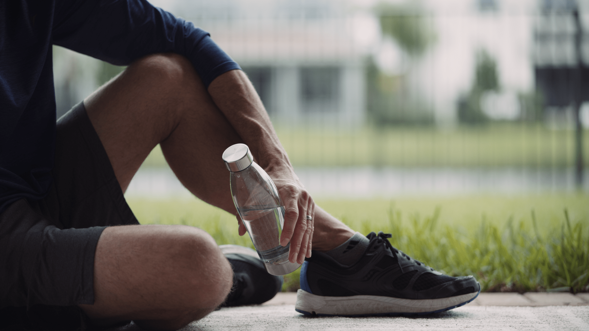 Side profile of man wearing black outfit sitting on the sidewalk holding a glass bottle of water