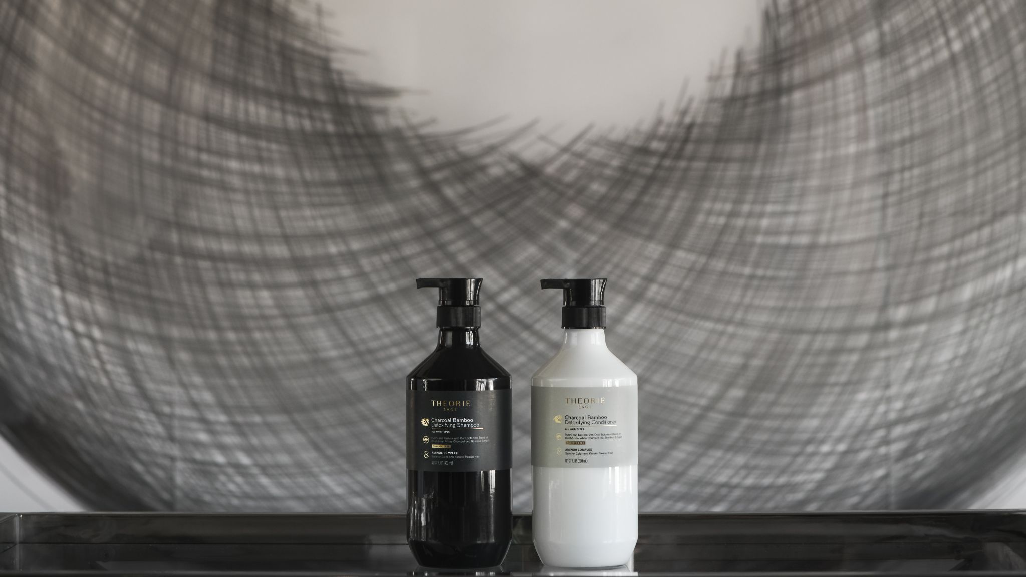 Theorie black detoxifying shampoo and white conditioner dispensers on a black table in front of a gray art form.