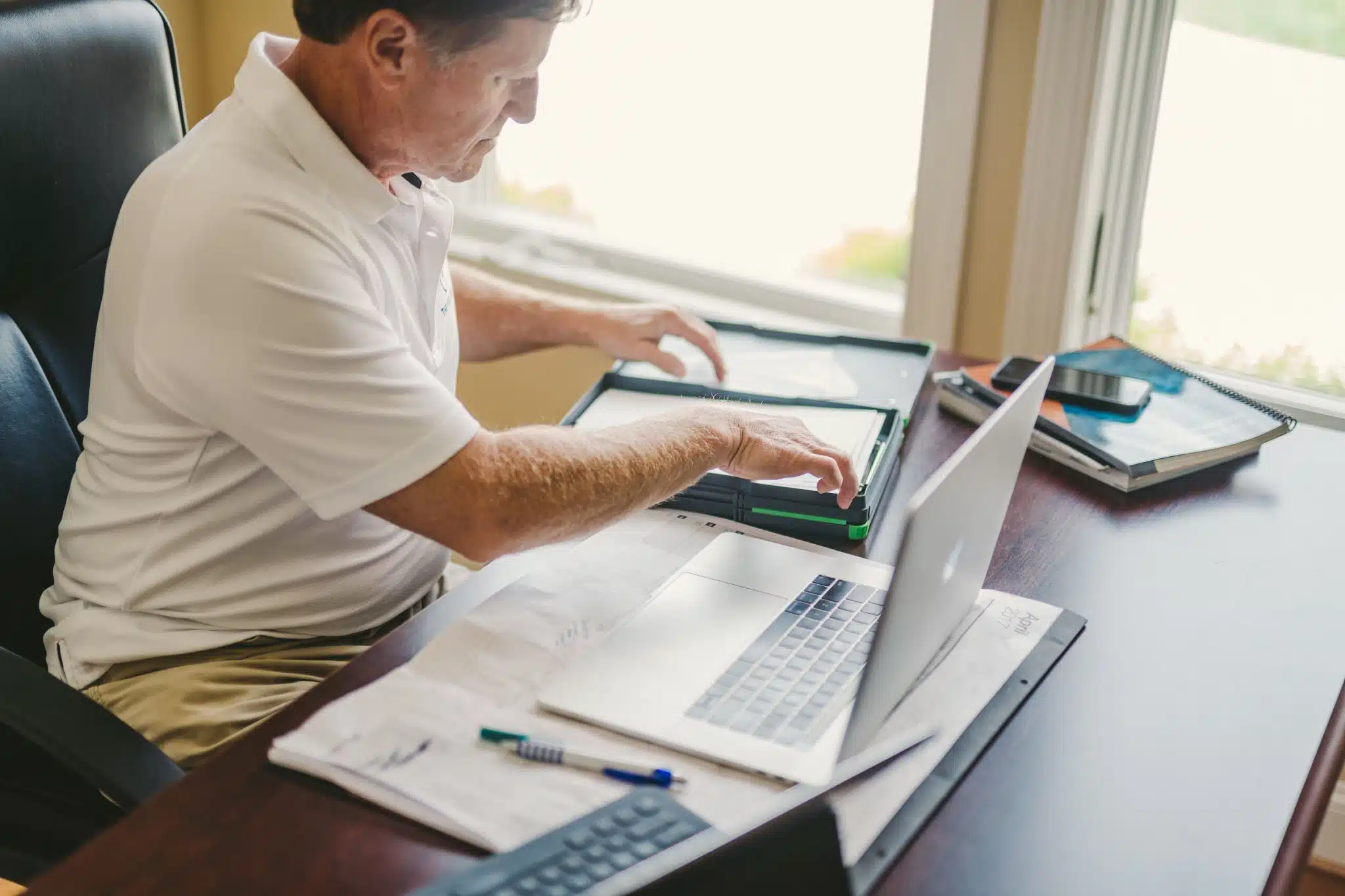 Older male in a white shirt and beige pants working at a desk with papers, laptops, books and calculator. He is sitting in a black, leather chair. Video Production Process