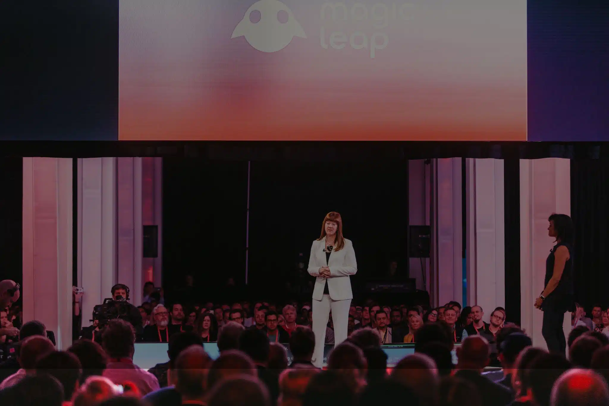 Event Photography Woman with red hair wearing a white pant suit with black top talking to audience from stage