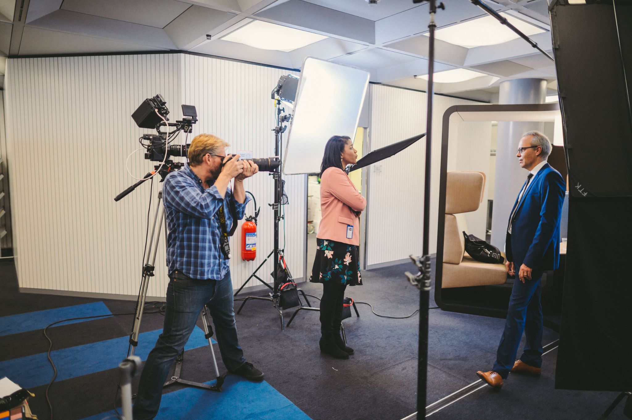 On a set with a plaid wearing photographer in blue jeans photographing a male in a blue suit and leather shoes being interviewed by a female in a flowery dress and salmon colored dress shirt.