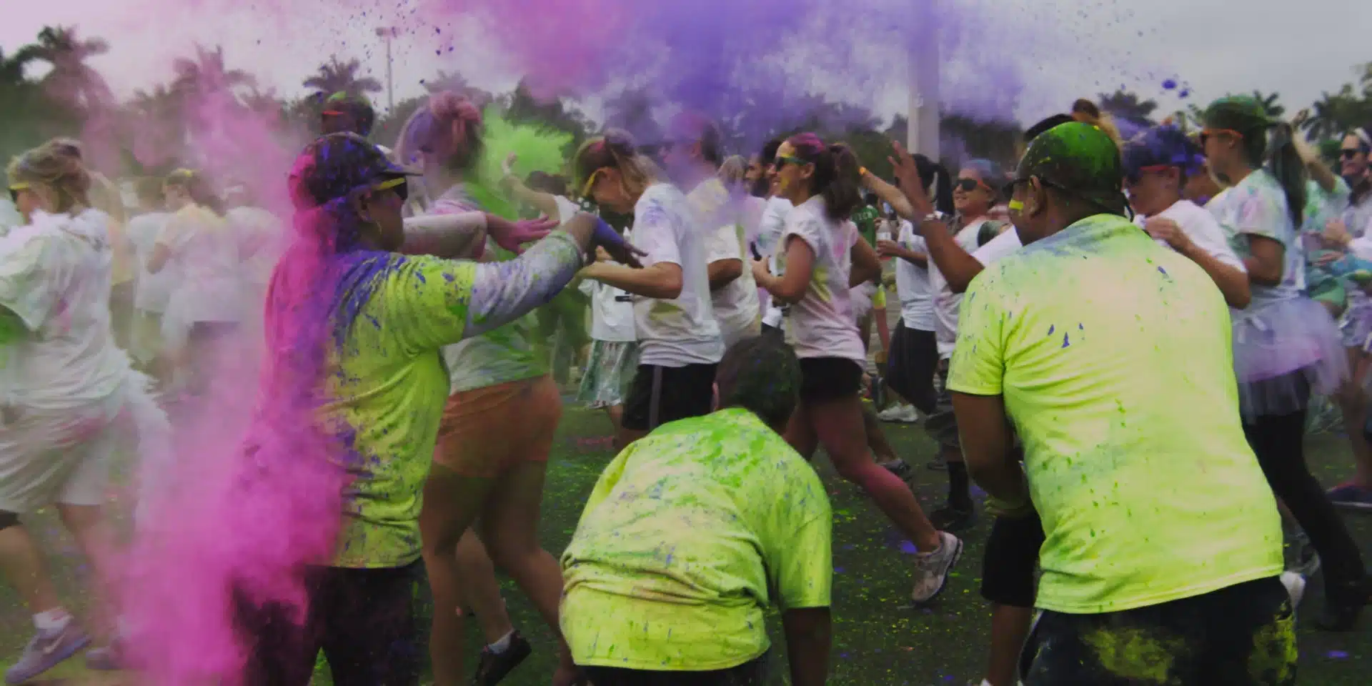 A bunch of people in colorful t-shirts releasing colored smoke for runners in white t-shirts running past.