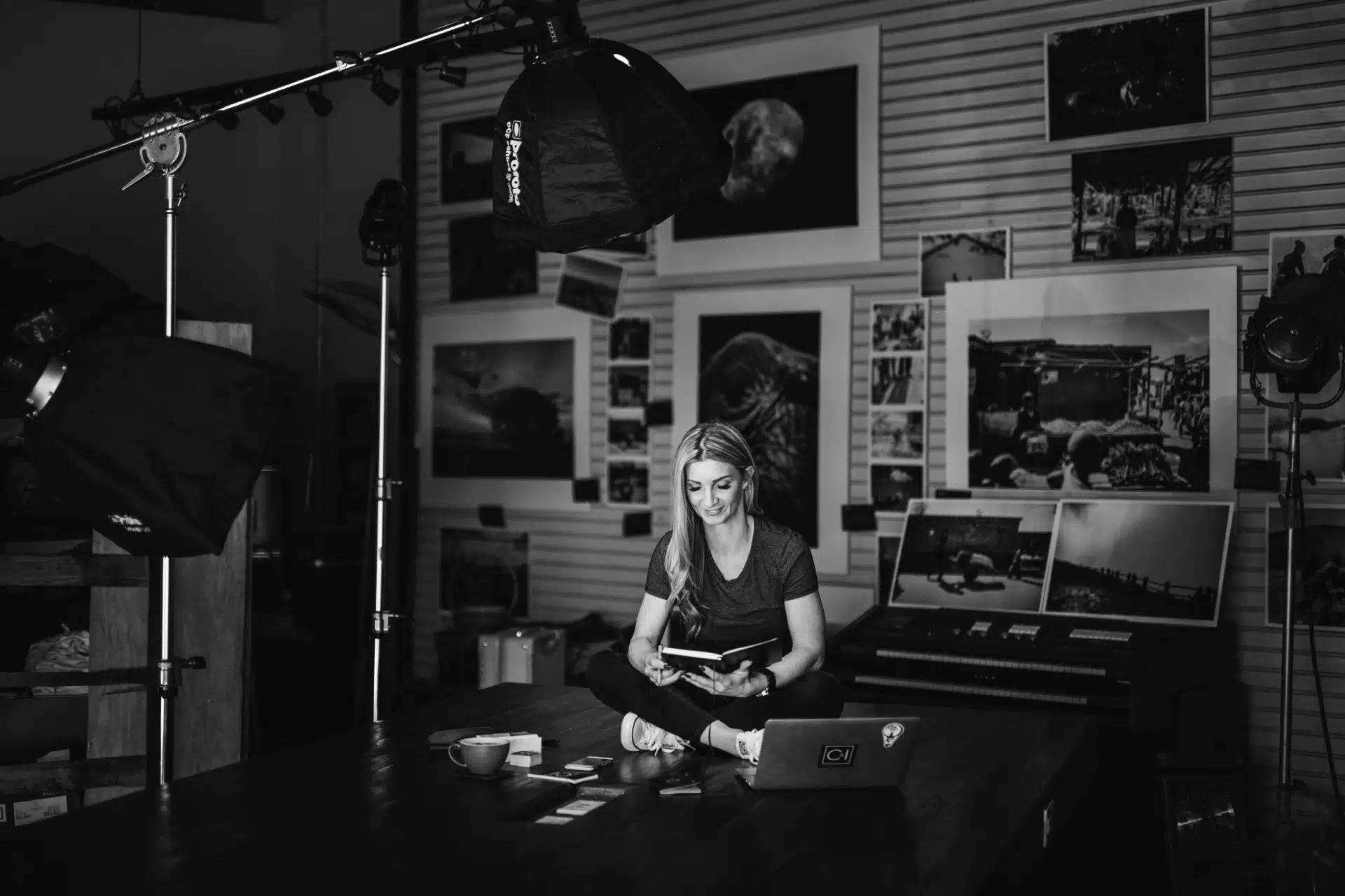 Black and white image of a woman with long hair sitting cross legged on the table with a laptop nearby reading a book. She is surrounded by art.