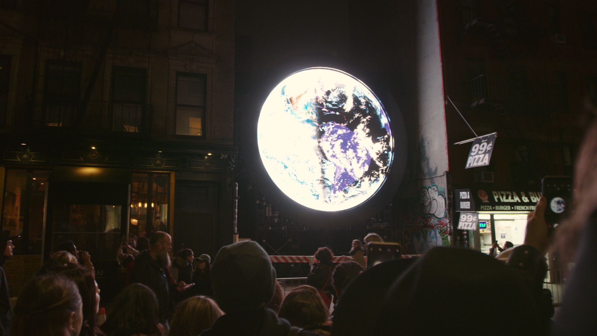 A crowd of people outside a building at night looking at art of a moon lit up in an alley.