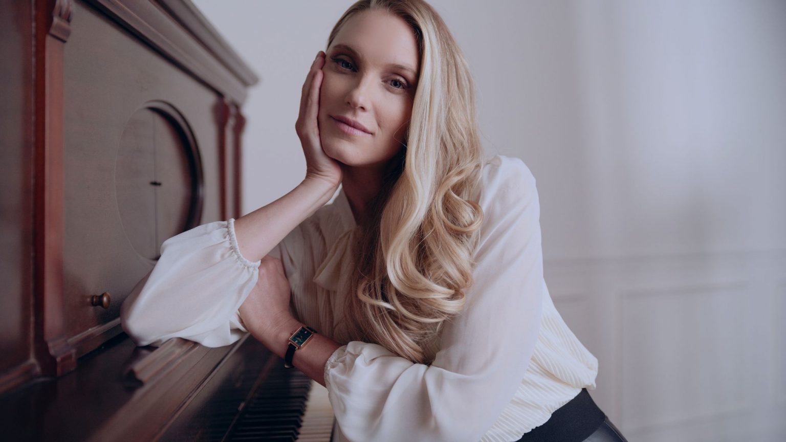 Woman With Long Blond Hair Wearing A White Top Posing For The Camera Leaning Against A Piano Sitting Down Wearing A Watch
