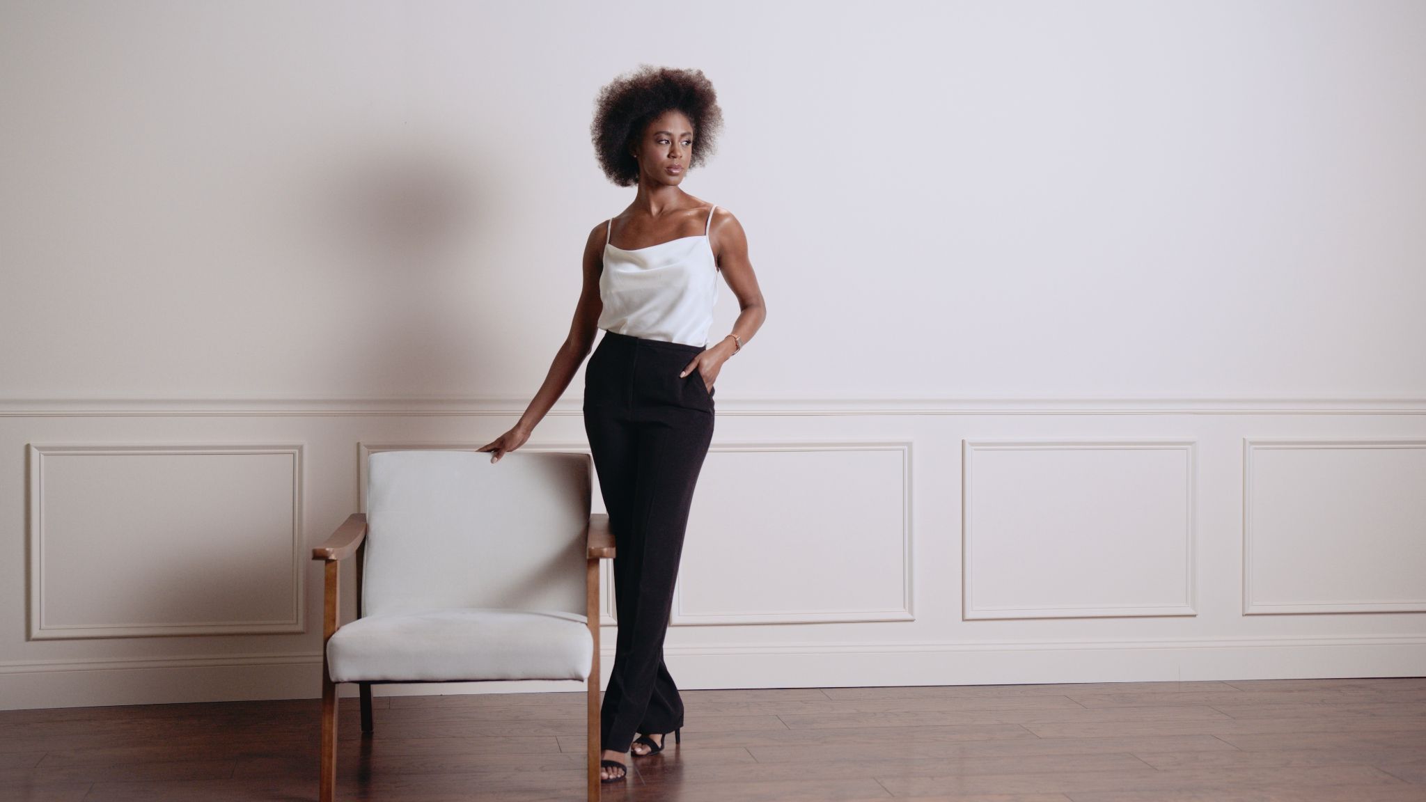 IU CI Studios Portfolio Lola Rose Lifestyle Products Sixty second Ad Still African American woman wearing white dress top, black pants and black heels posing for the camera holding the backing of a white chair next to her