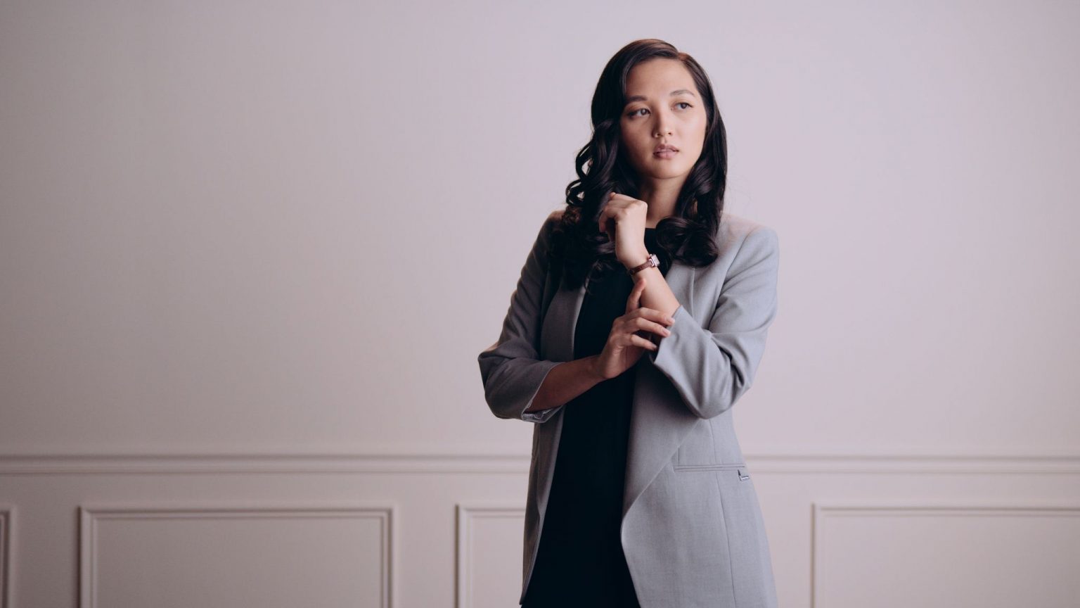 Asian Woman With Long Black Hair Wearing A Black Top With Gray Dress Jacket Posing For Camera Wearing A Watch