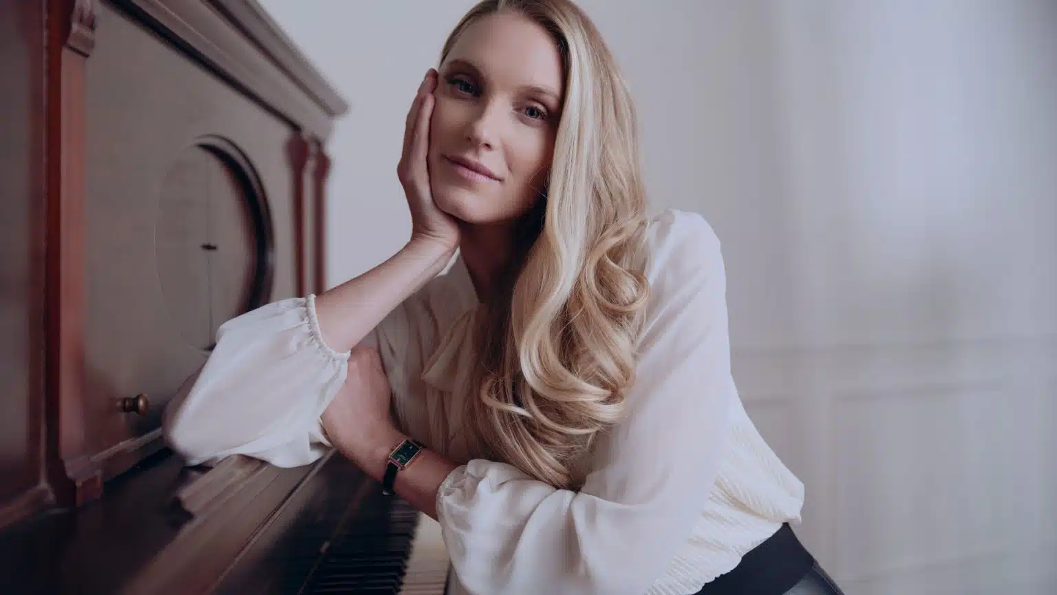 Woman with long blond hair wearing a white top posing for the camera leaning against a piano sitting down wearing a watch