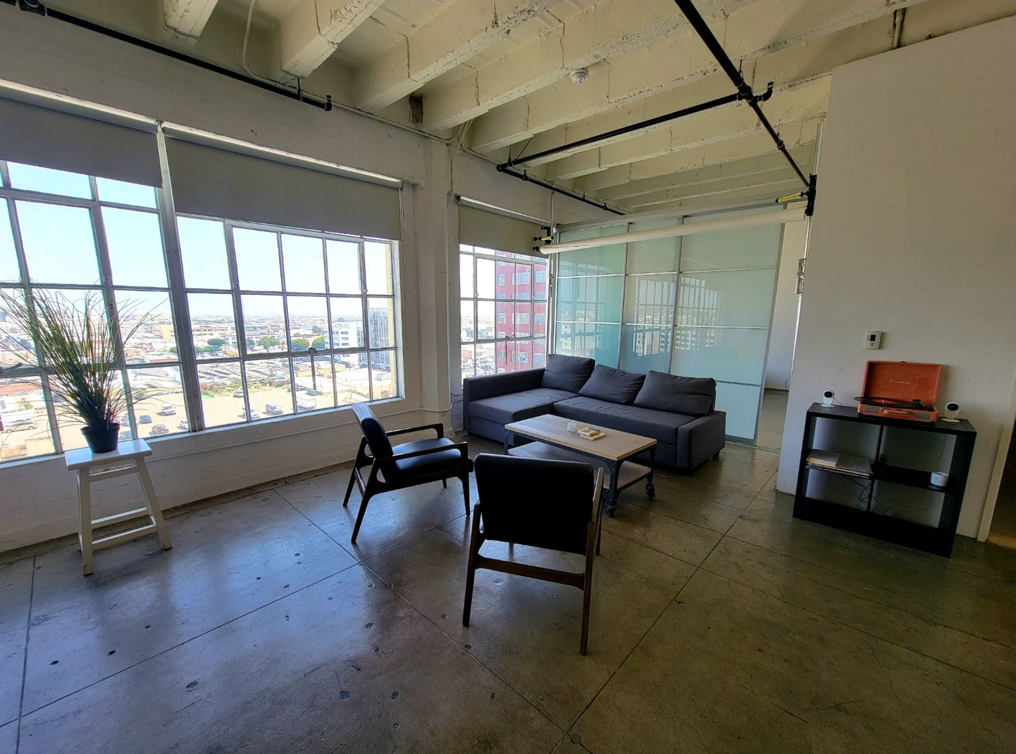 Room overlooking city with two slate gray chairs and a wraparound couch. There is a white table with a plant on it. There is also another table with an old orange vinyl record player.