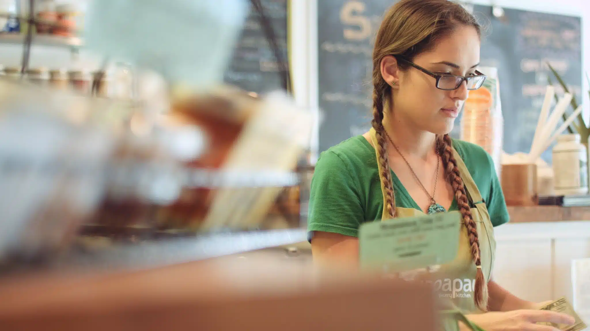 Woman with blond braids wearing glasses and green t shirt and apron handling money at a business