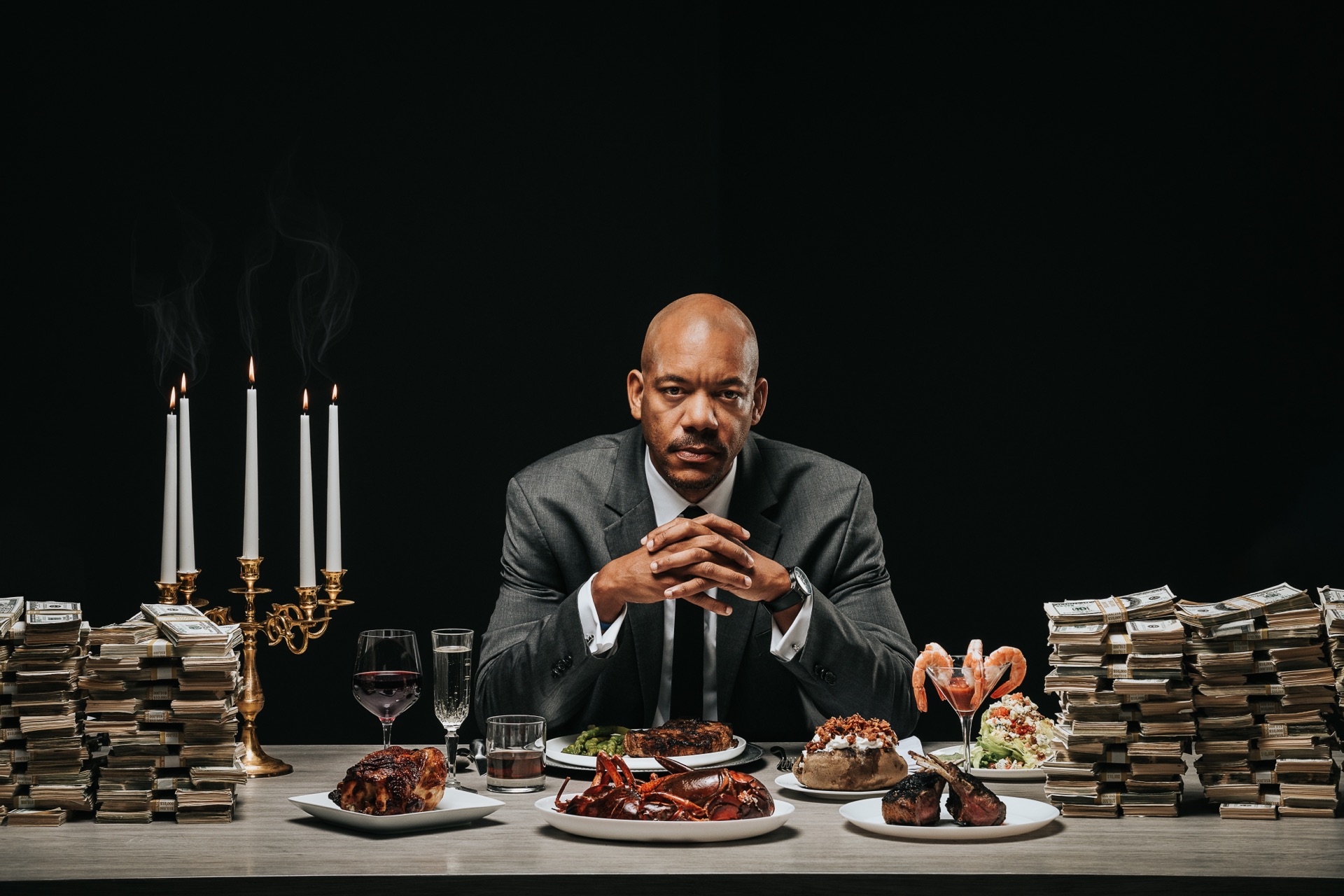 African American male at a table surrounded by exotic foods, cash and a candelabra with lit candles.