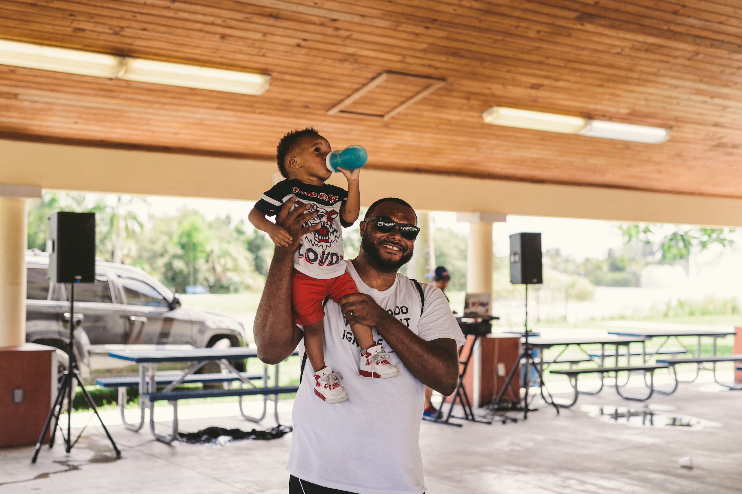 KES African American man wearing a white t shirt holding a toddler on his shoulder who is drinking from a sippy cup posing for the camera from inside a pavilion
